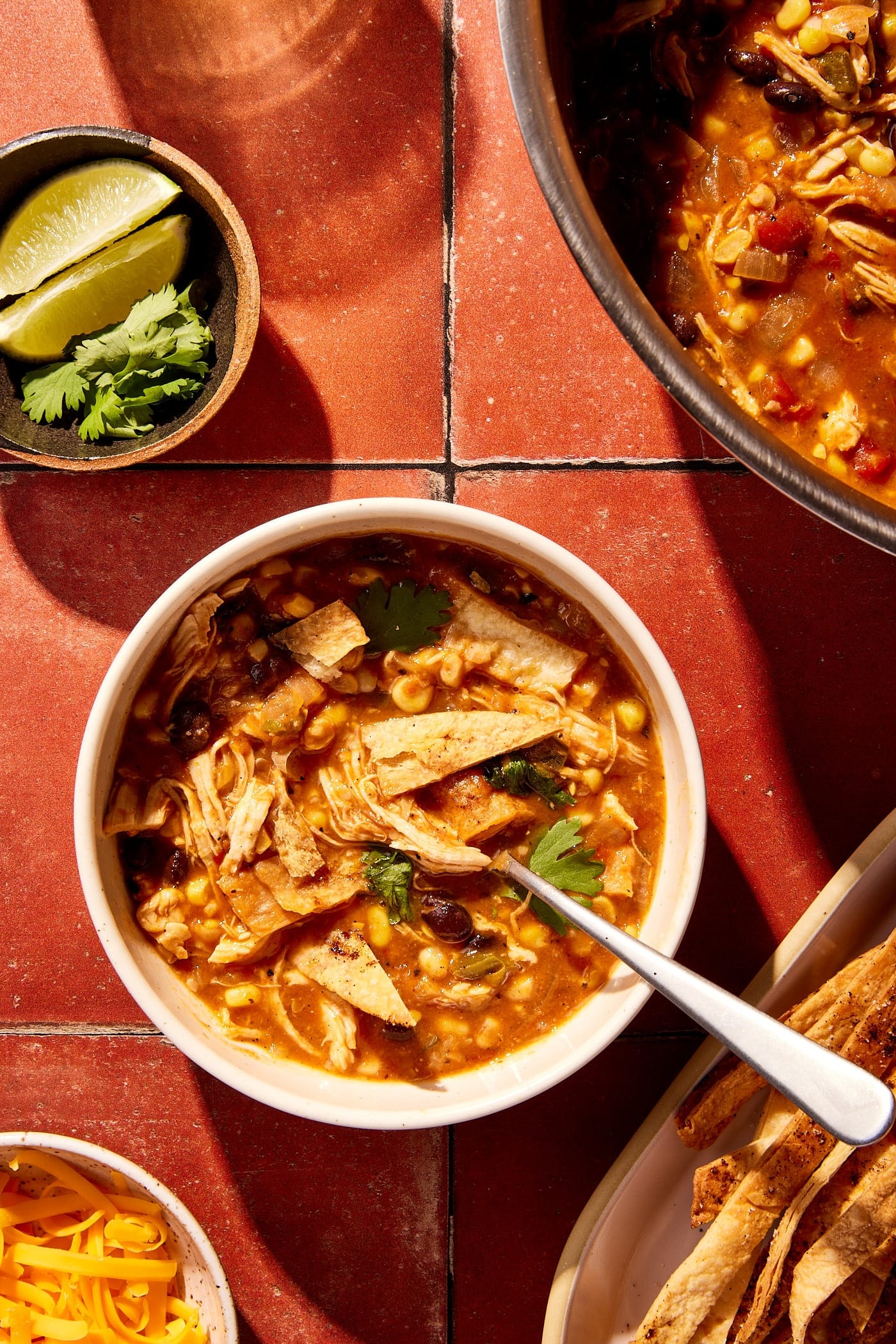 Red tile surface with bowl of chicken tortilla soup with spoon in bowl along with lots of toppings all around.
