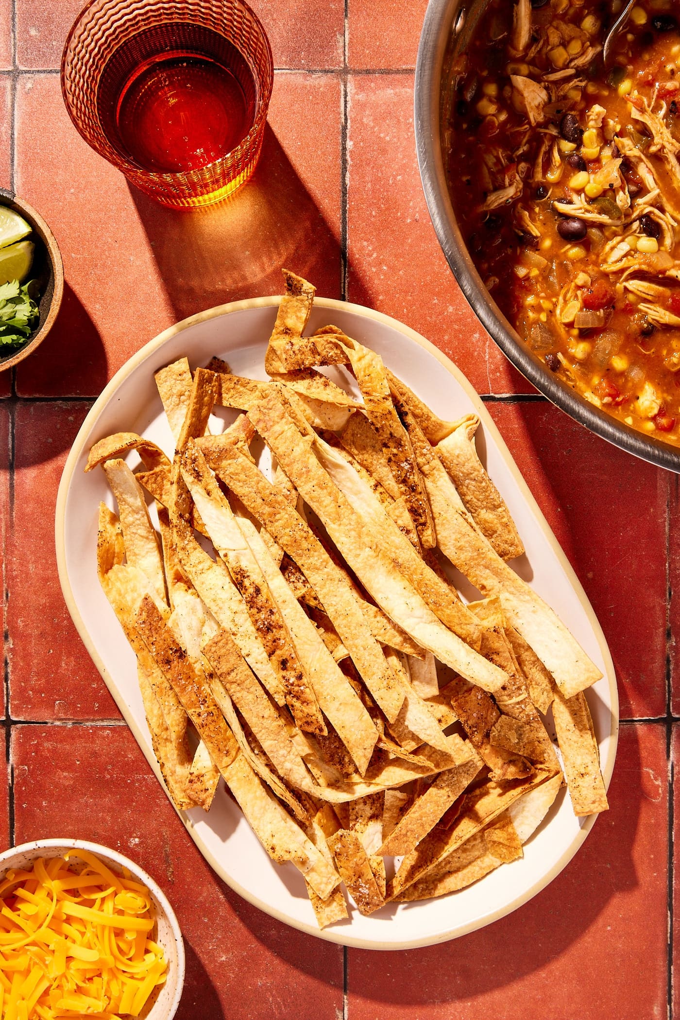 White platter filled with baked crispy tortilla strips on red tile surface with pot filled with chicken tortilla soup to the side.