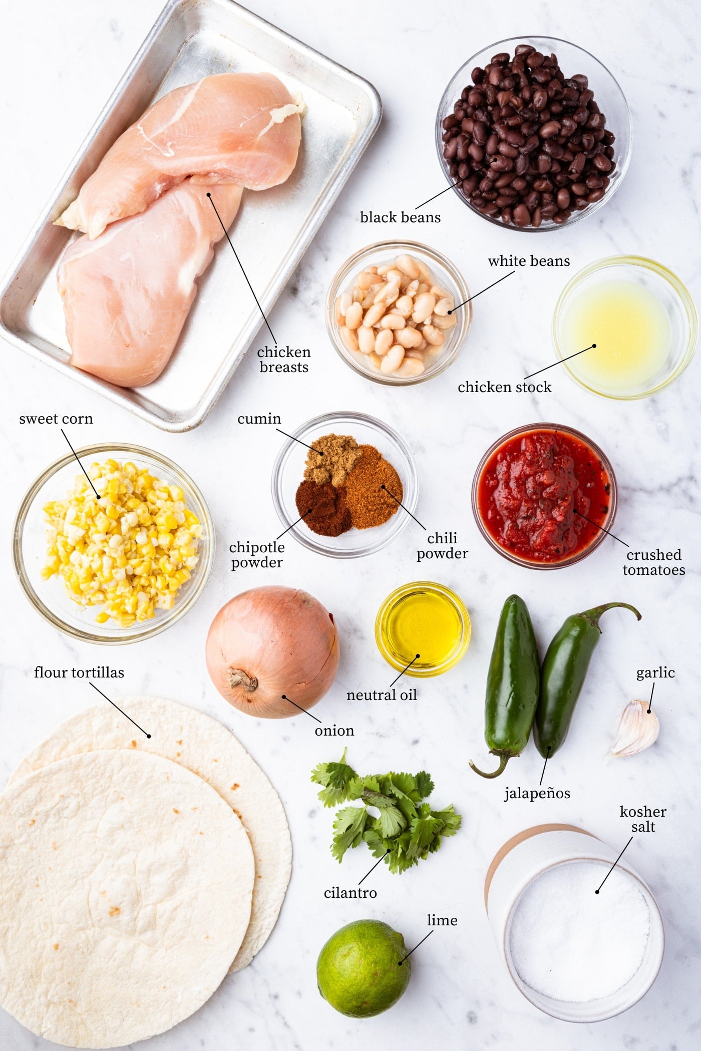 White marble surface filled with all ingredients needed to make chicken tortilla soup including raw chicken, corn, spices, and more.