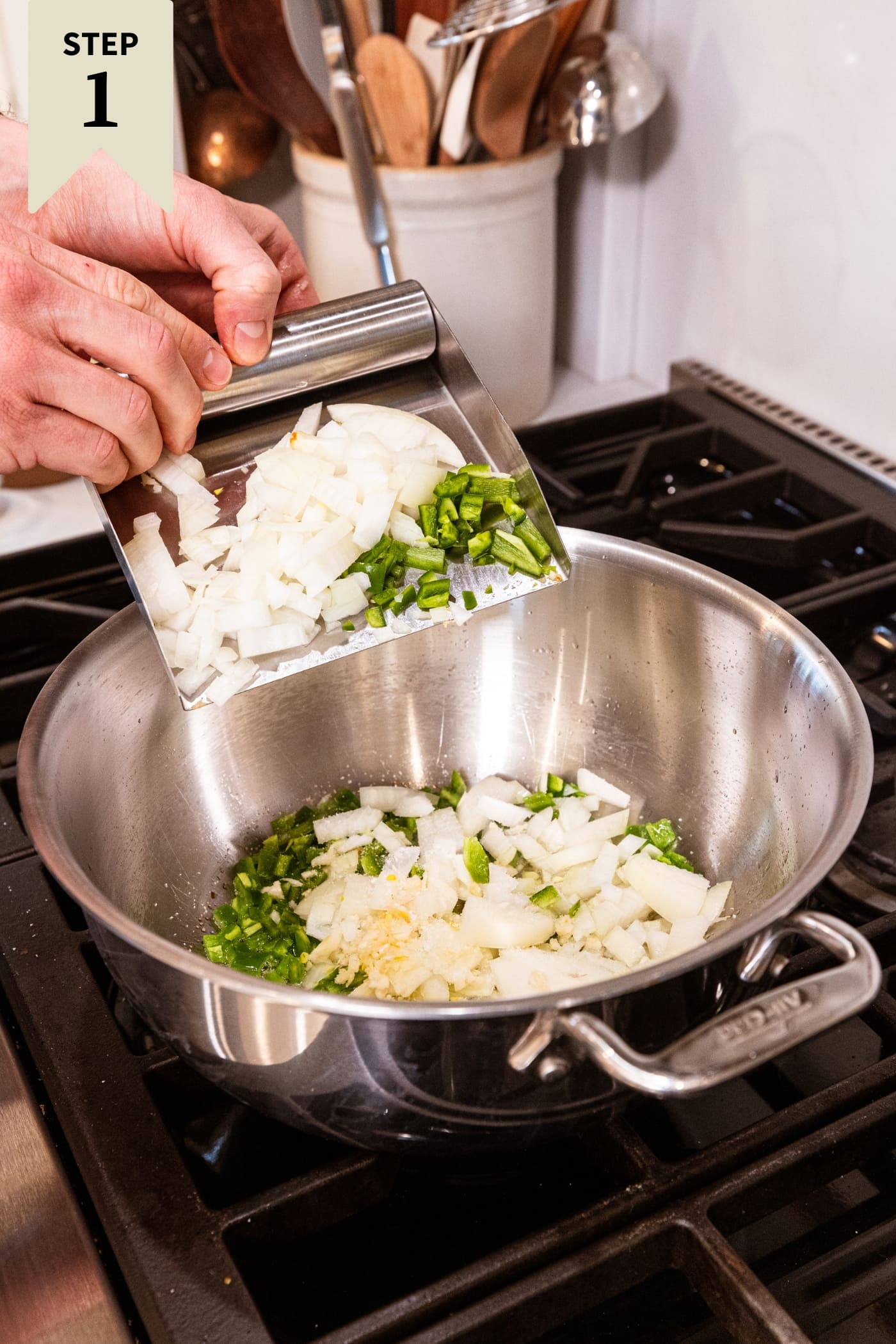 Hand holding metal scraper adding diced onion, jalapeño, and garlic to a large stockpot on the heat.