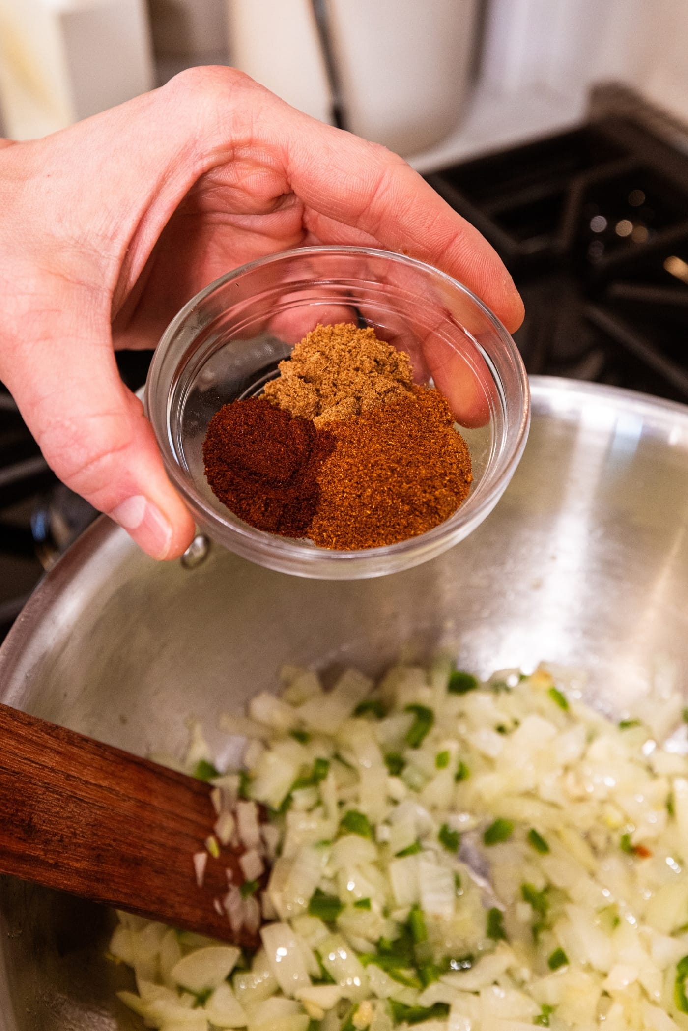Hand holding small glass bowl filled with three different spices about to pour on top of diced onion mixture.