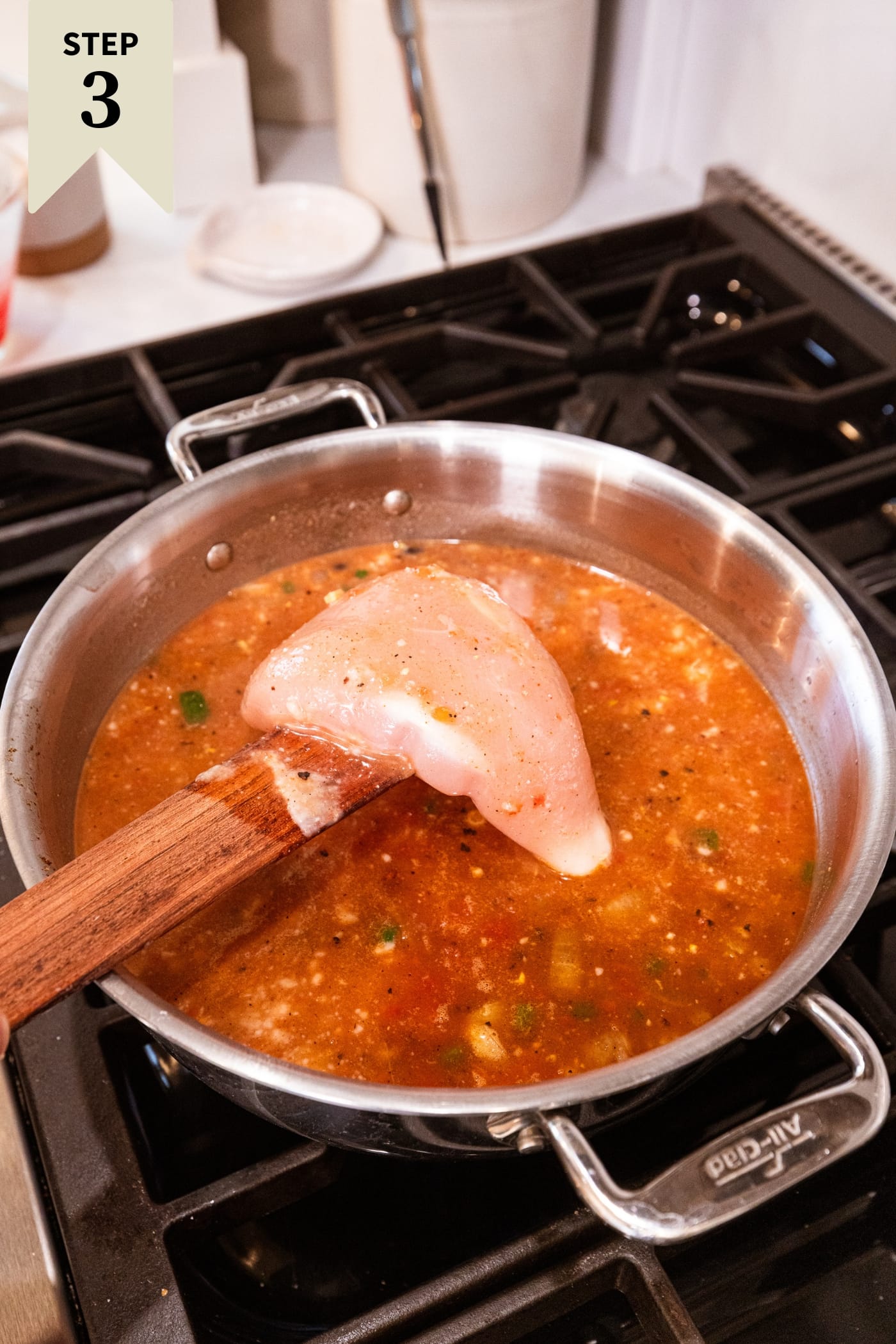 Wooden spatula about to place raw chicken breast into soup mixture on the stovetop.