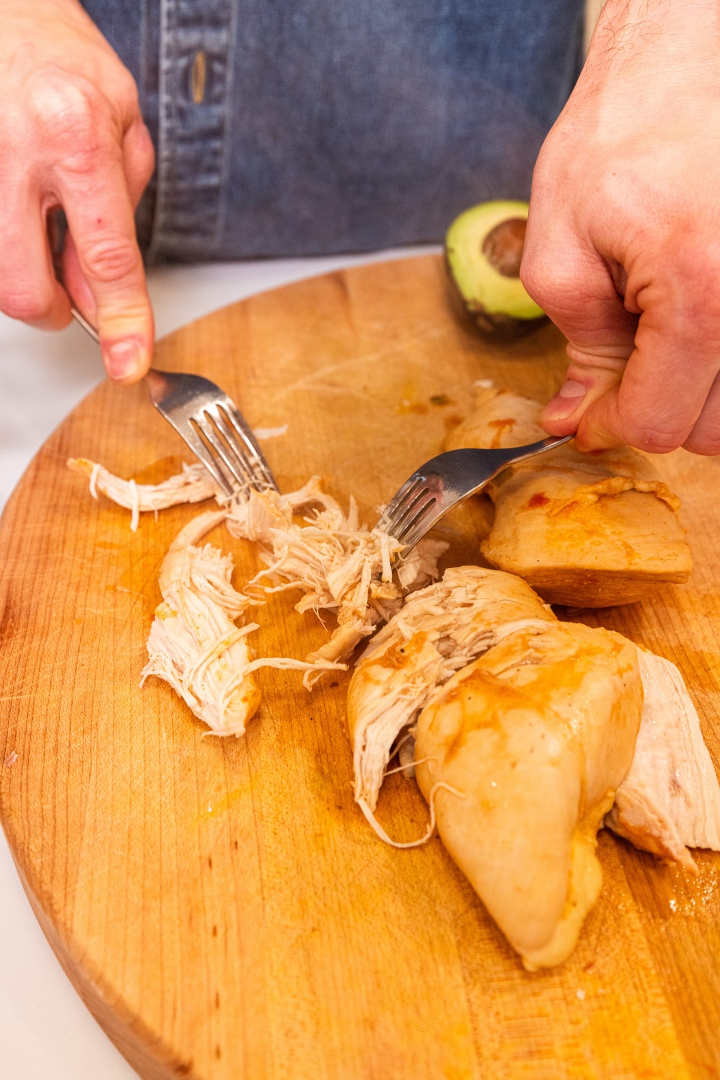 Two forks being used to shred cooked chicken breast into pieces on wooden cutting board.