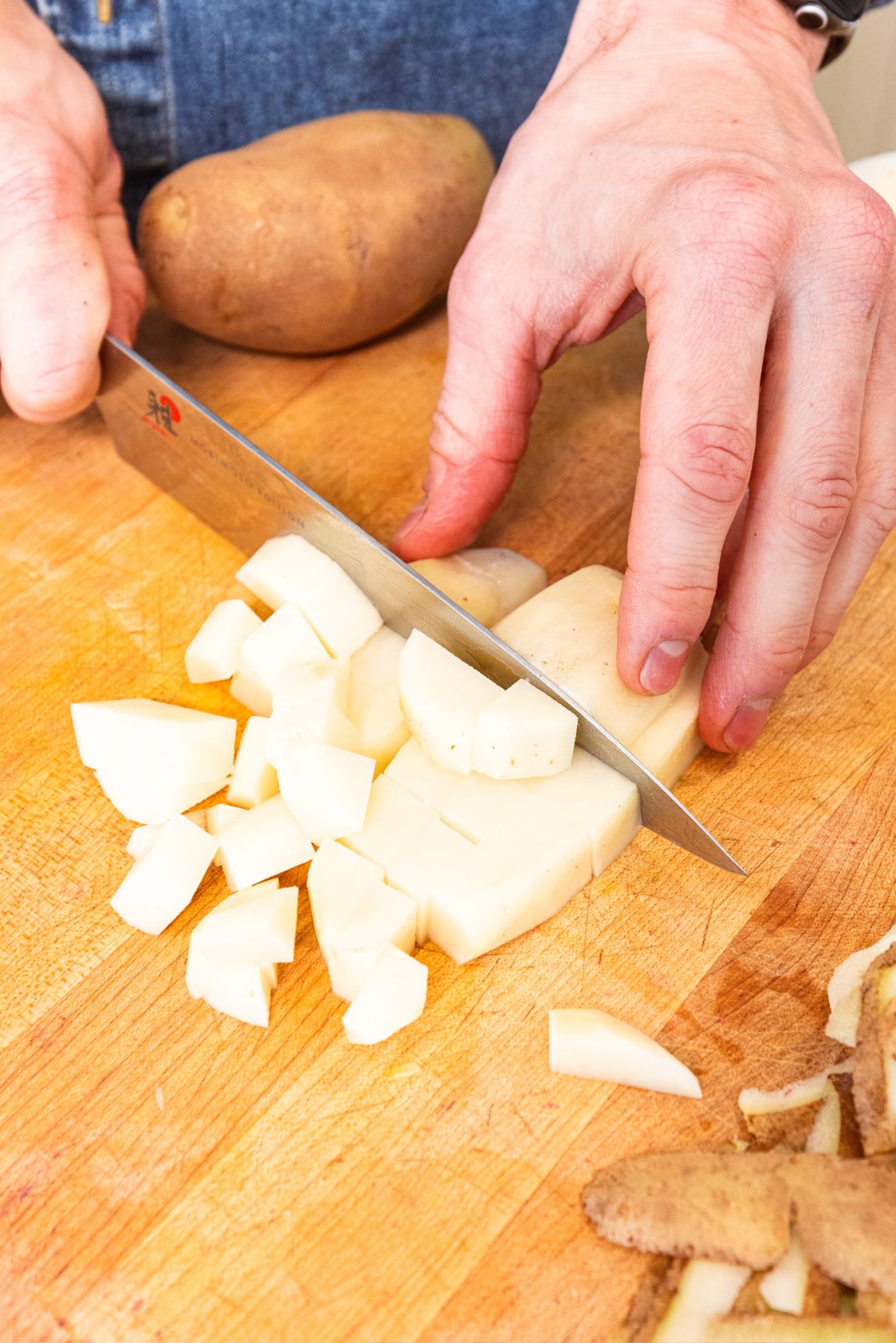 Hand using a knife to slice a potato into diced cubes on a wooden cutting board.
