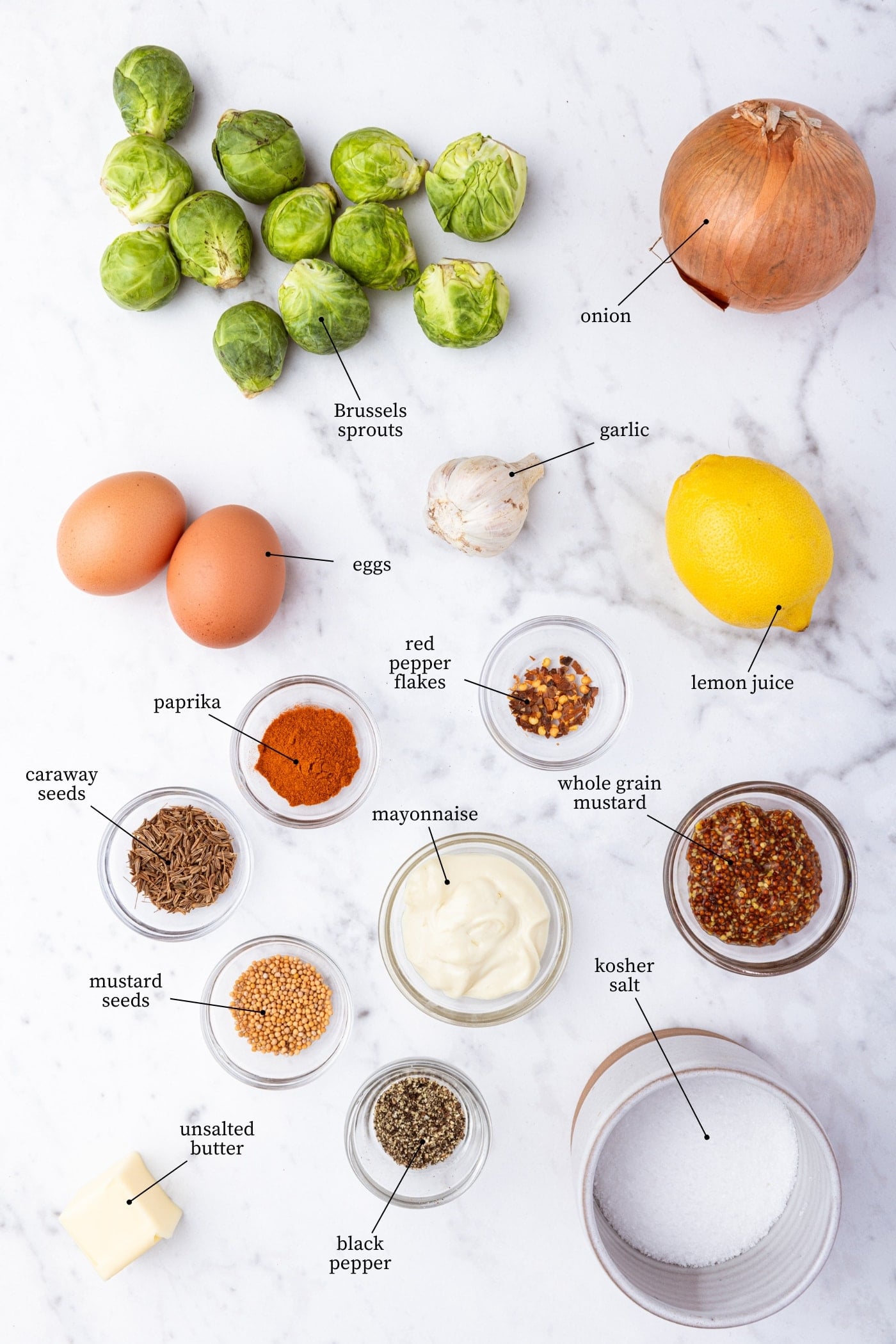 White marble surface filled with all ingredients needed to prepare Brussels sprouts hash including vegetables, eggs, spices, onion, and more.