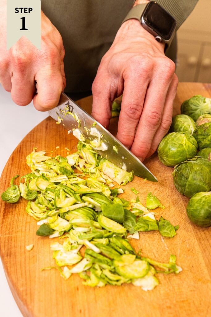 Hand using knife to chop Brussels sprouts into thin strips on a wooden cutting board.
