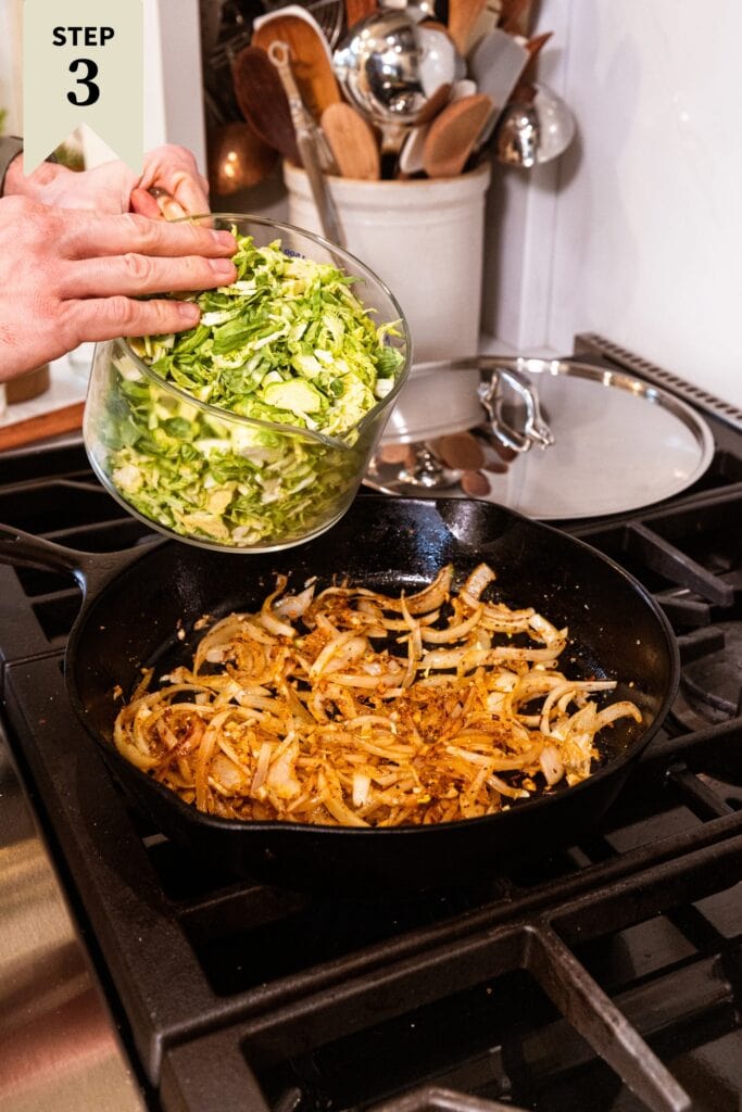 Black cast iron skillet filled with brown cooked onions with hand about to pour in chopped Brussels sprouts from a glass measuring cup.