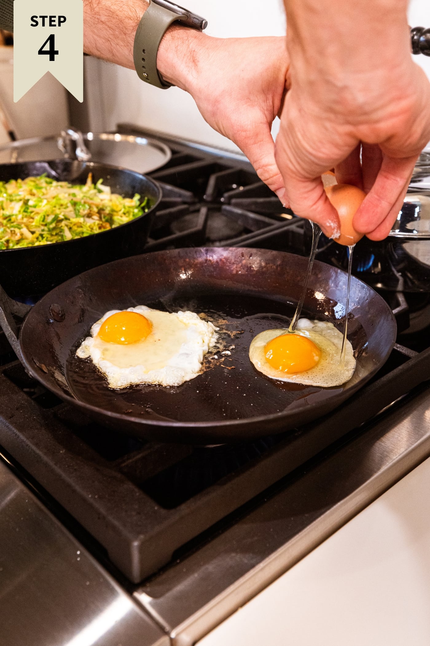 Hand opening a egg from the shell into a large cast iron skillet with an egg already cooking.