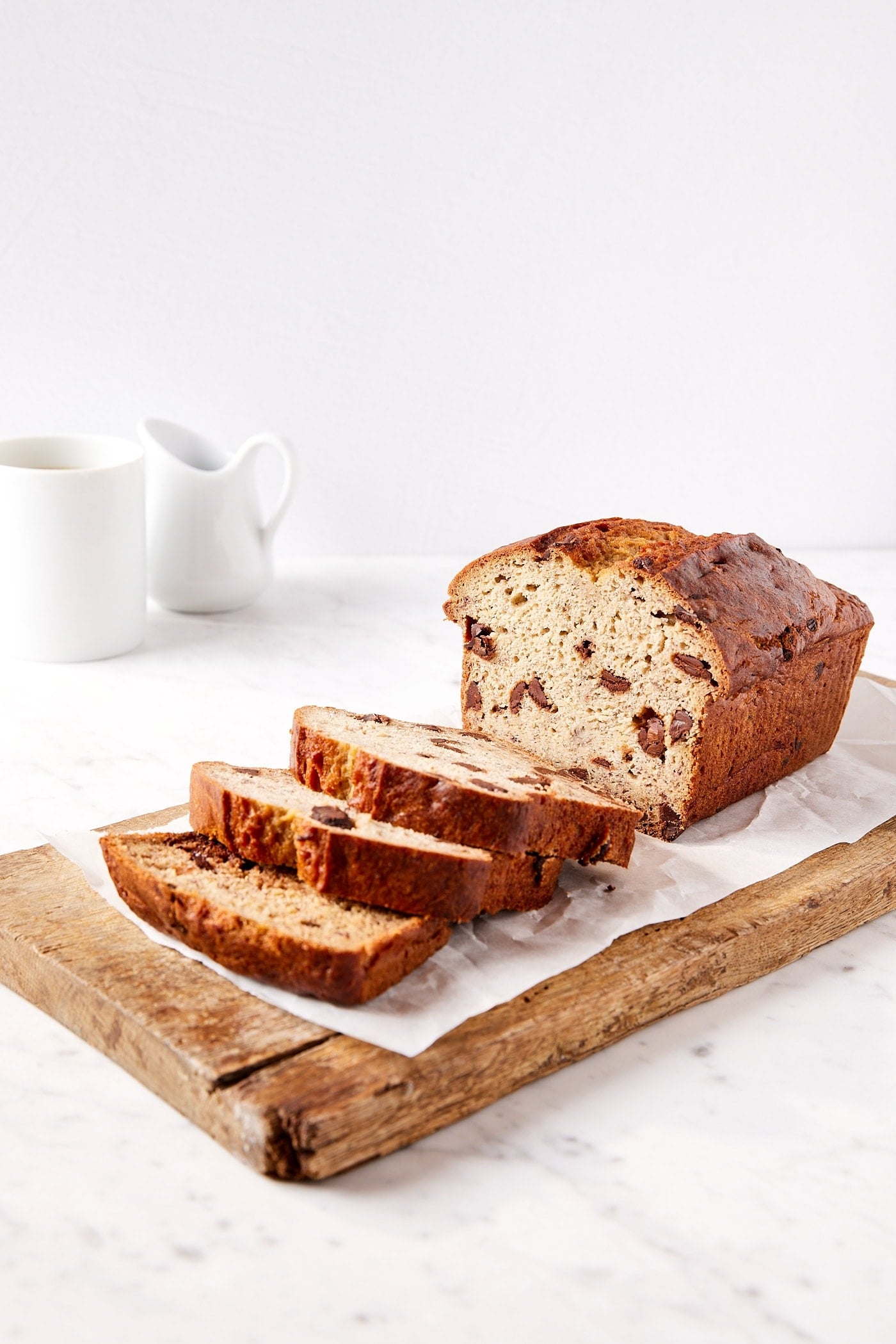 Loaf of chocolate chip banana bread resting on wooden cutting board on white marble surface with white coffee cup.