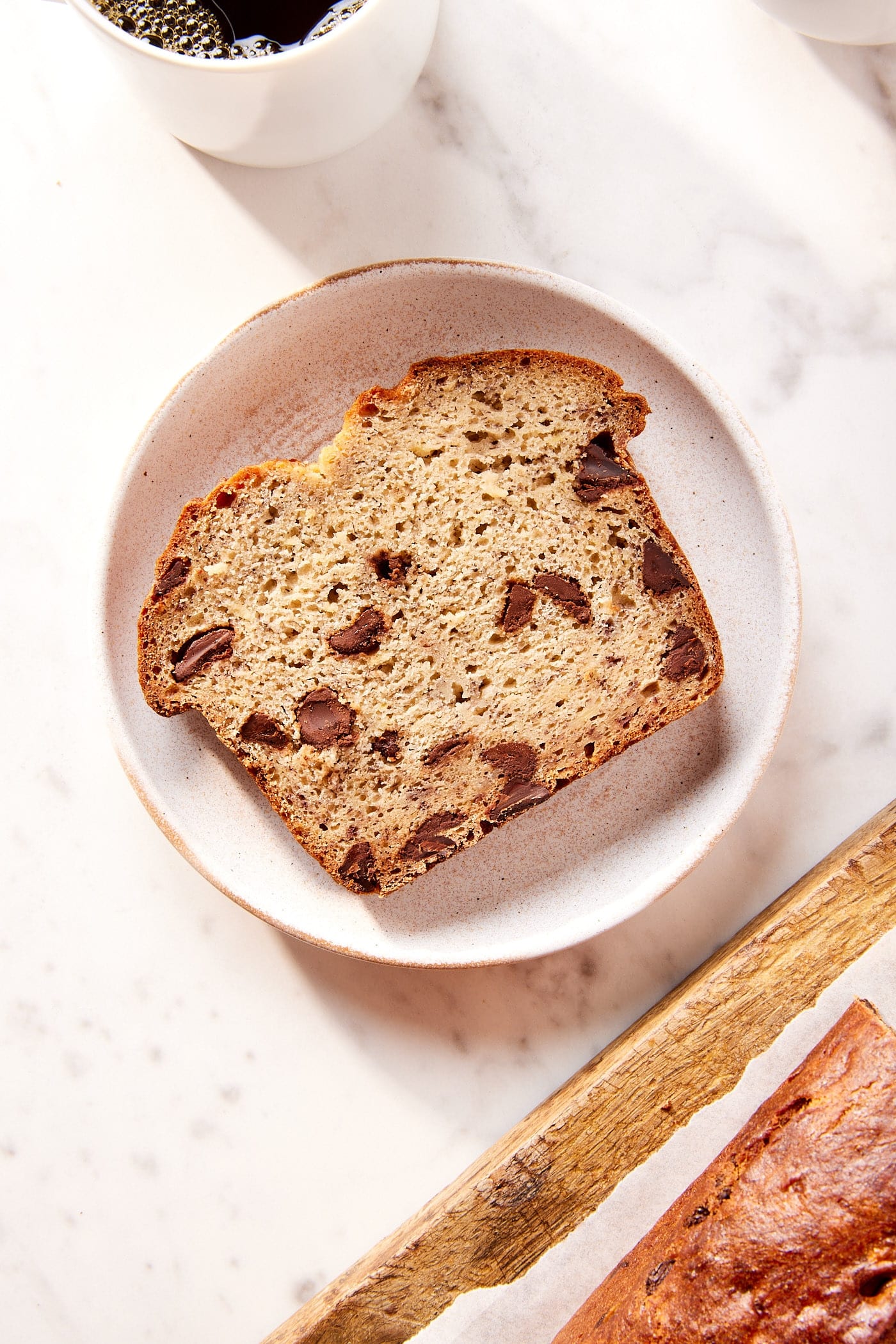 Slice of chocolate chip banana bread resting on a plate with chunks of chocolate showing with cup of coffee to the side.