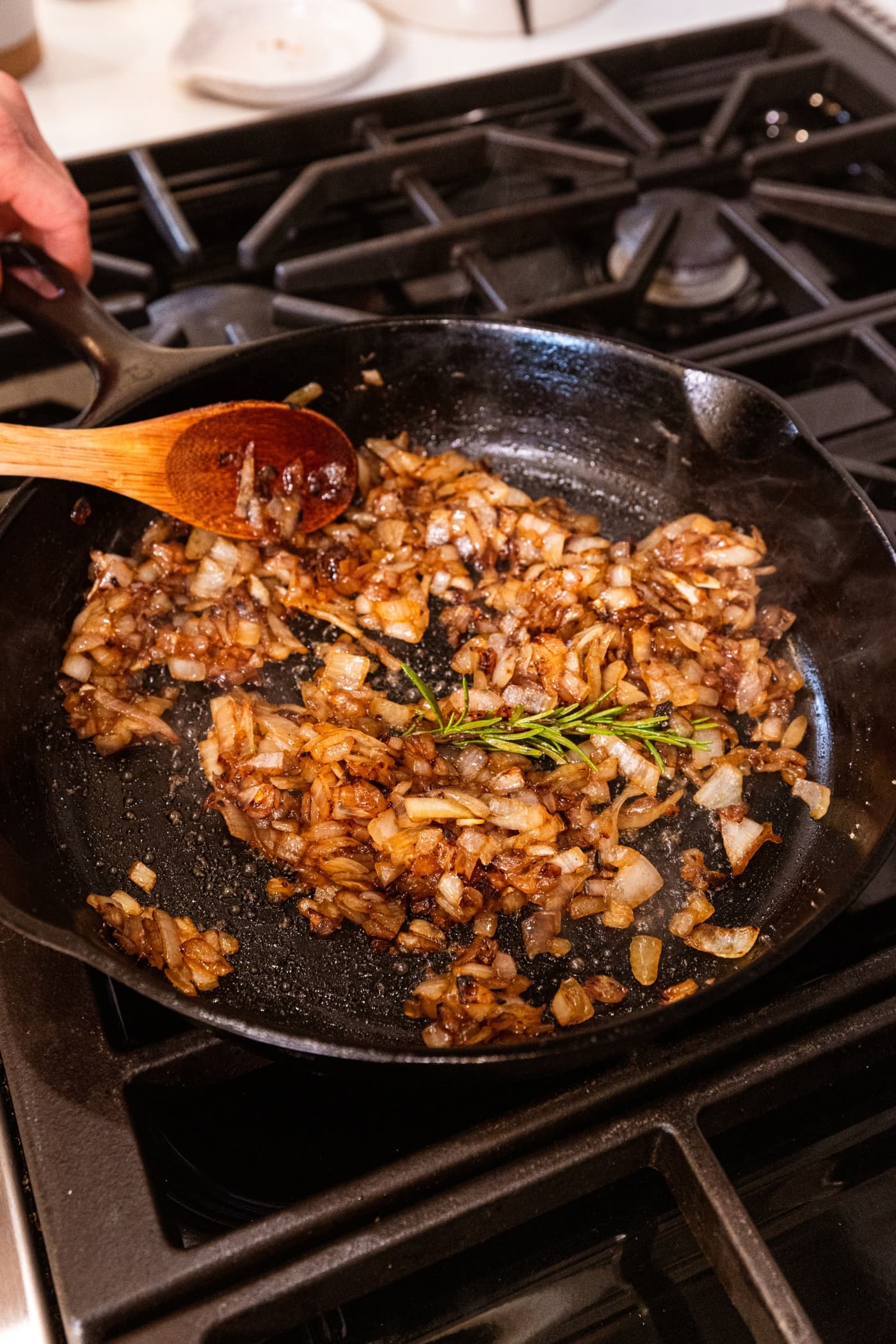 Wooden spoon scraping together browning onions in a black cast iron skillet with a sprig of rosemary.