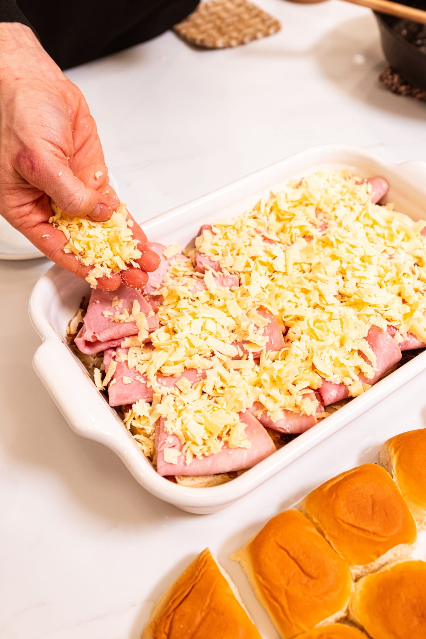 Shredded cheese being sprinkled all over the top of slices of ham in a white baking pan.