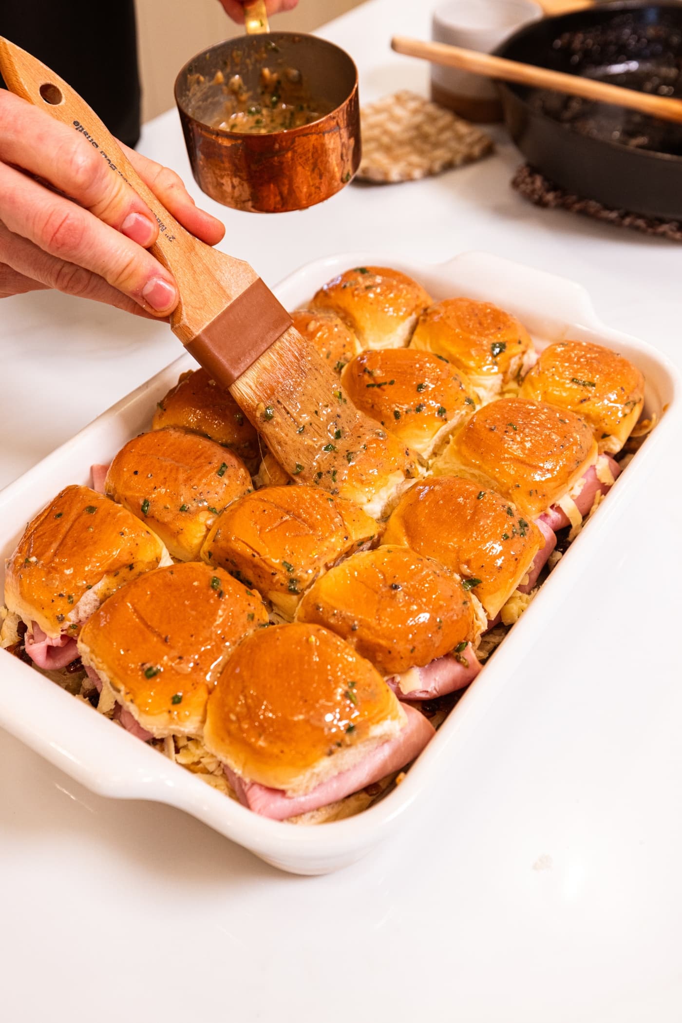 Brush brushing melted butter mixture with herbs over the top of sweet rolls in a white baking pan.