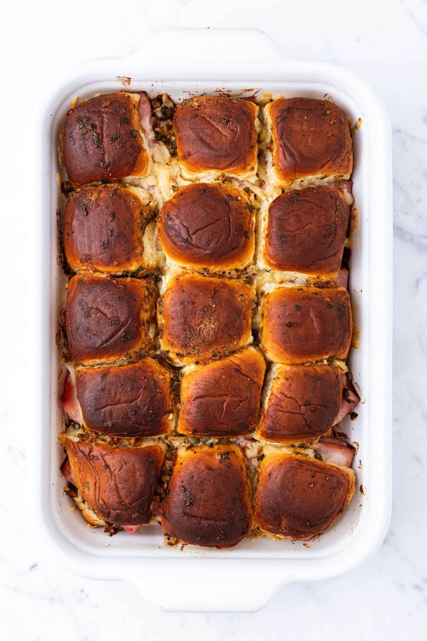 White baking dish filled with baked ham and onion jam sliders on a marble surface right after coming out of the oven.