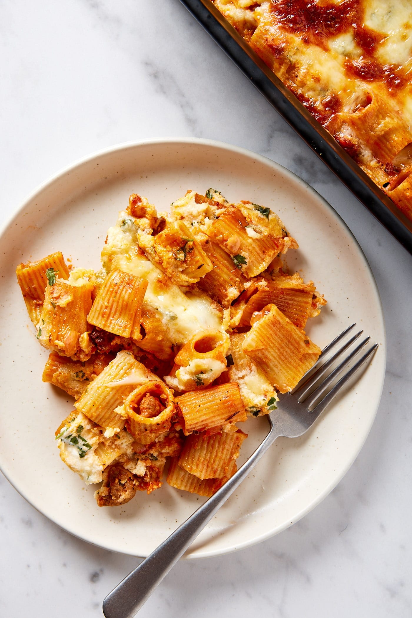 White plate filled with serving of lasagna casserole with fork resting on the plate with rest of baking dish filled with lasagna.
