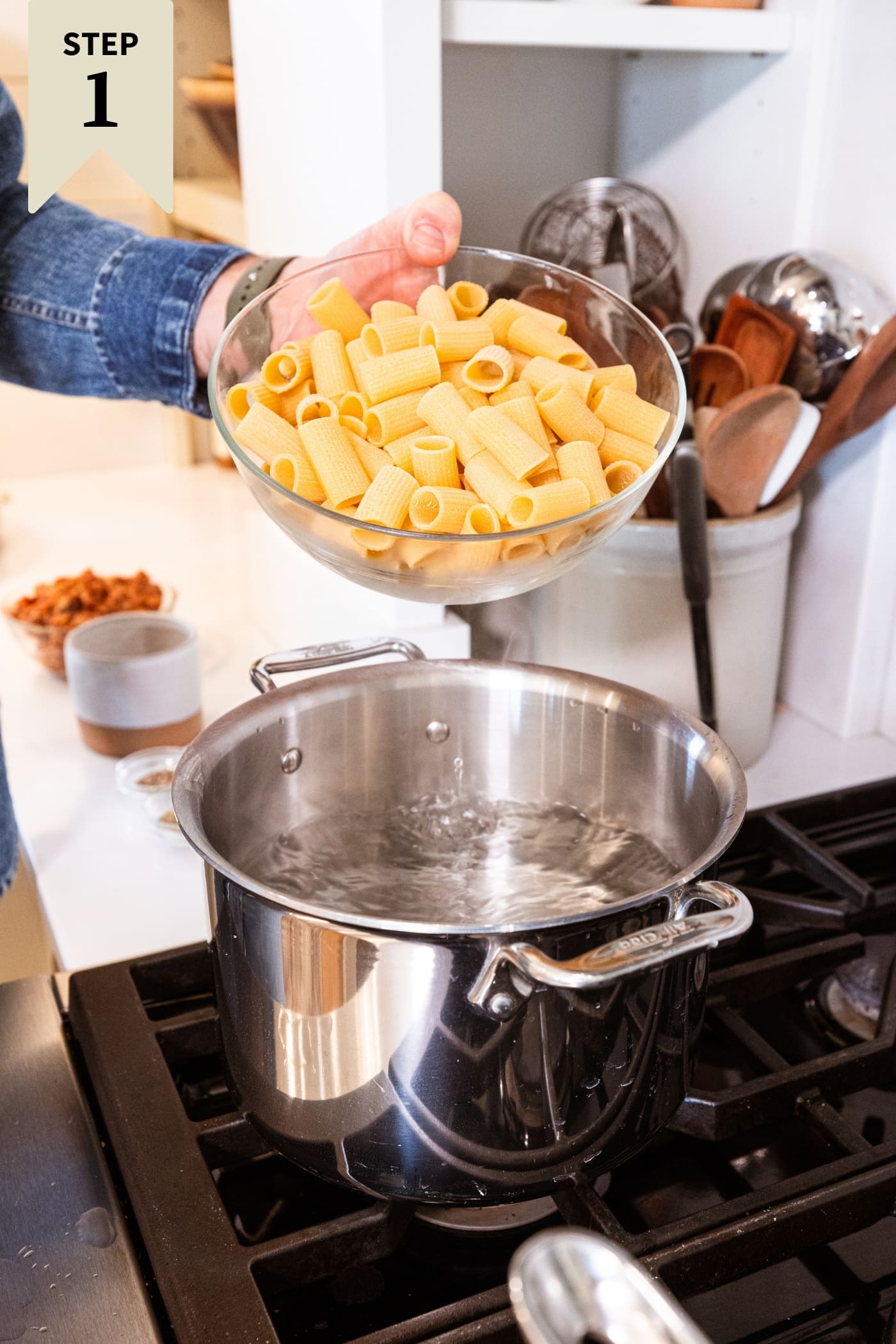 Hand holding glass bowl filled with pasta about to pour it into a large stockpot on the stove filled with boiling water.