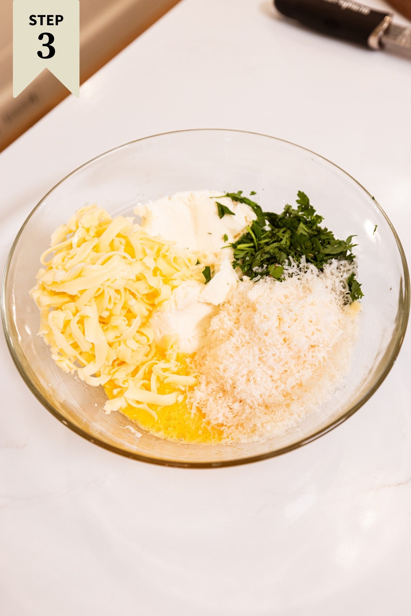 Glass bowl on white surface filled with ingredients needed for a homemade ricotta filling including parsley and cheese.
