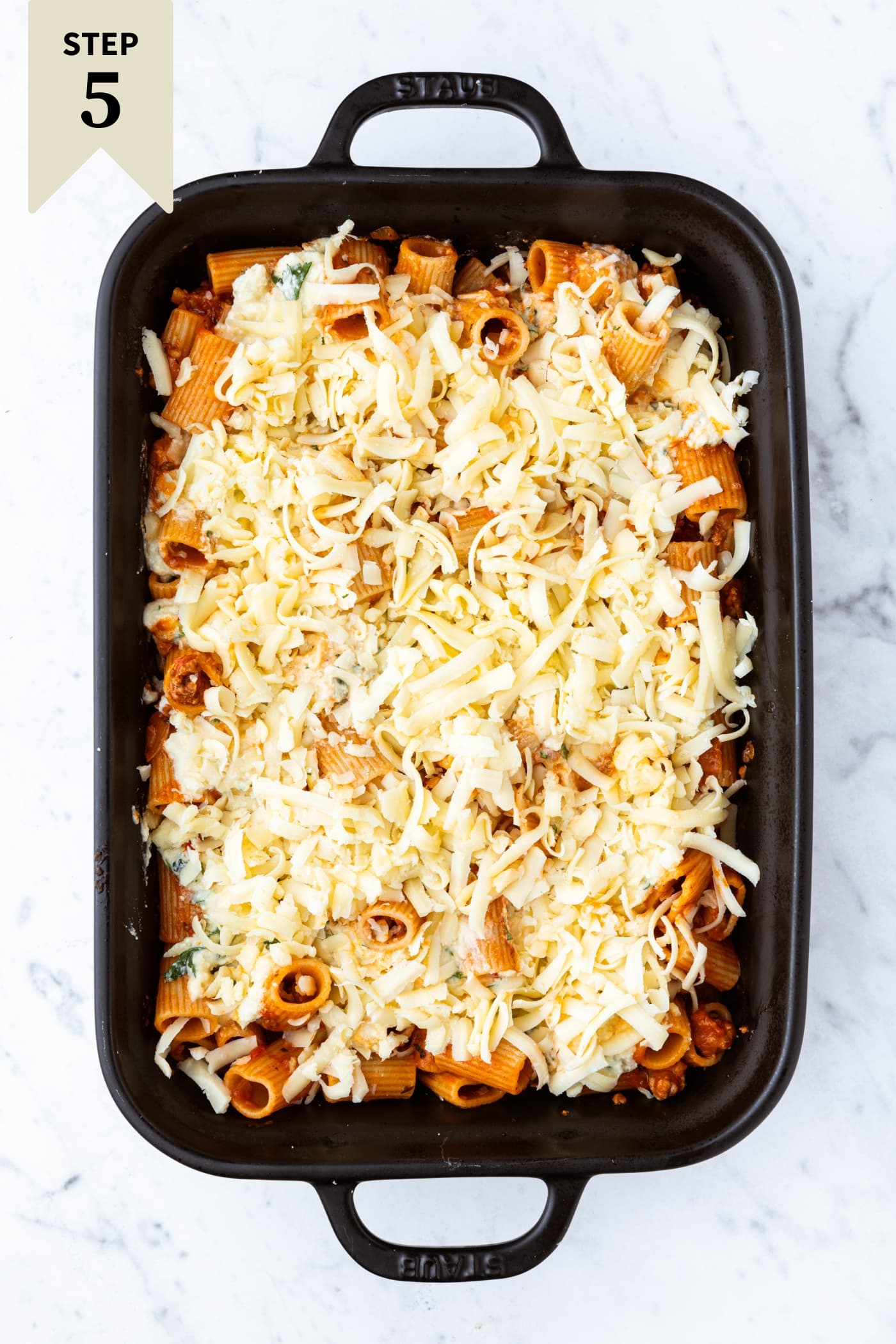 Black rectangular baking dish filled with unbaked lasagna casserole on white marble surface.