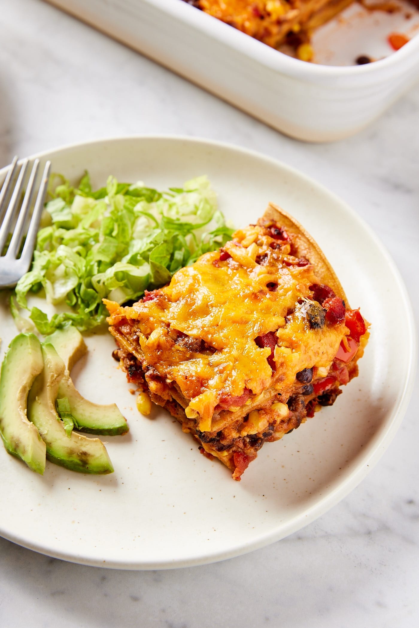 White plate with slice of loaded taco squares casserole with lettuce and slices of avocado with fork to the side all on white marble surface.