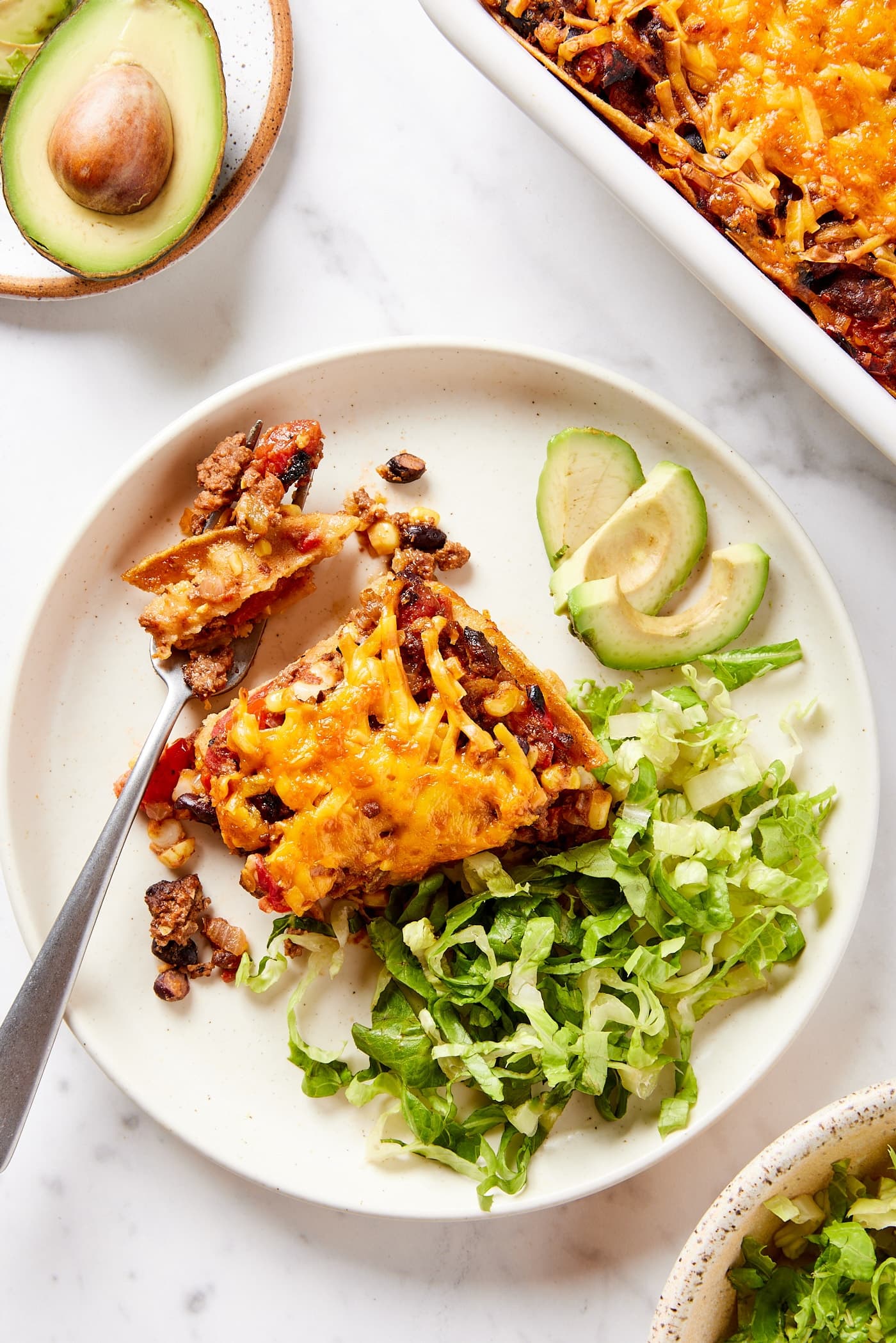 White marble surface with white plate with square of taco squares casserole with piece cut off with fork about ready to eat.