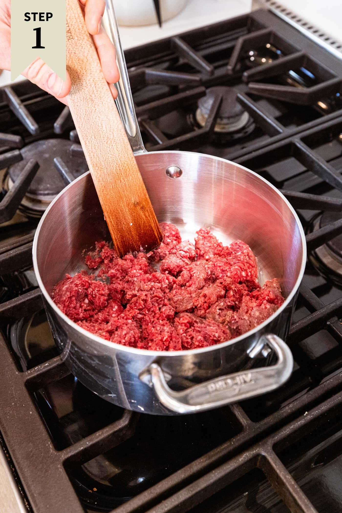 Metal saucepan on the stovetop with raw ground beef before being cooked with a wooden spatula to the back.