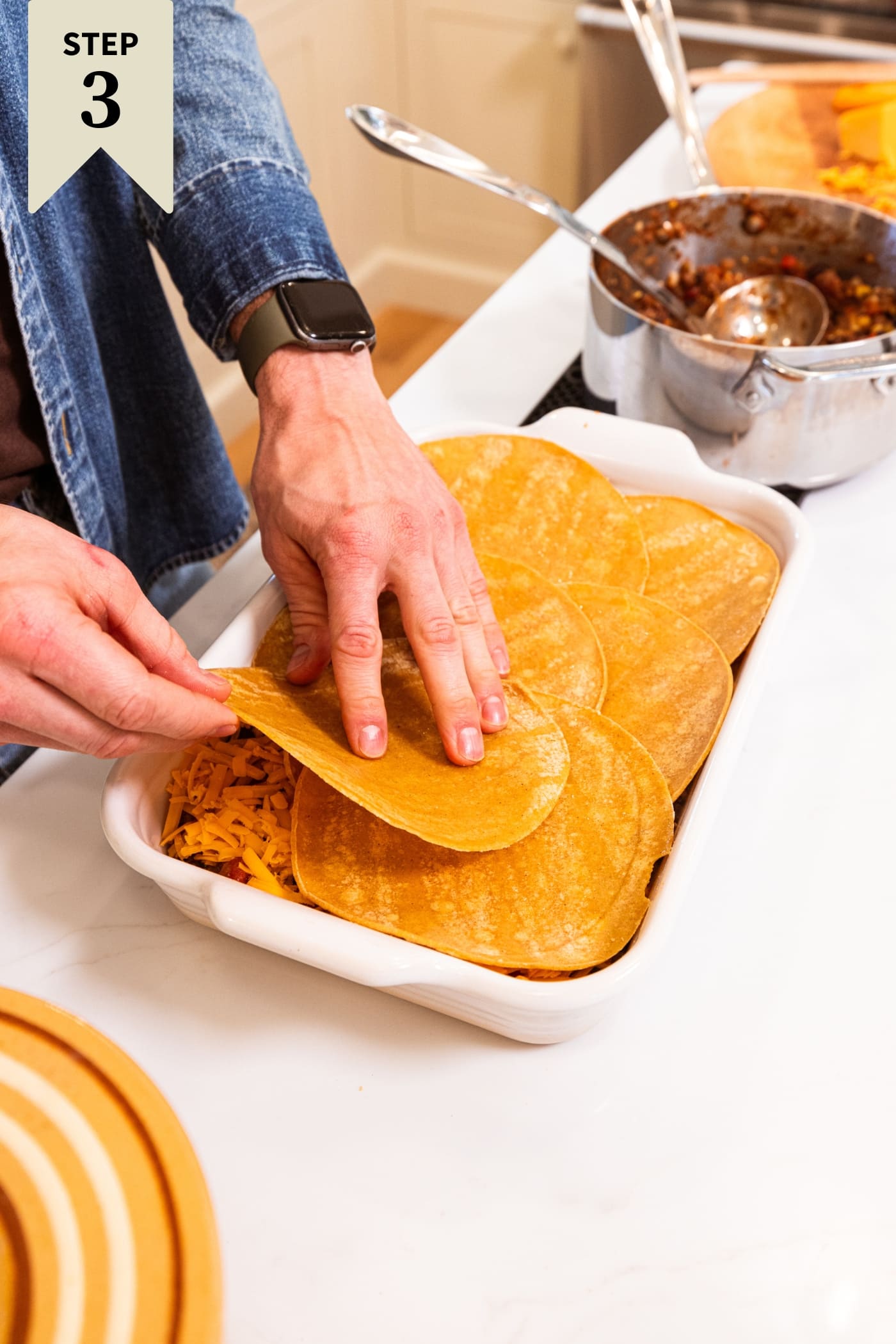 Hand placing corn tortillas in a white baking dish with ground beef mixture underneath.