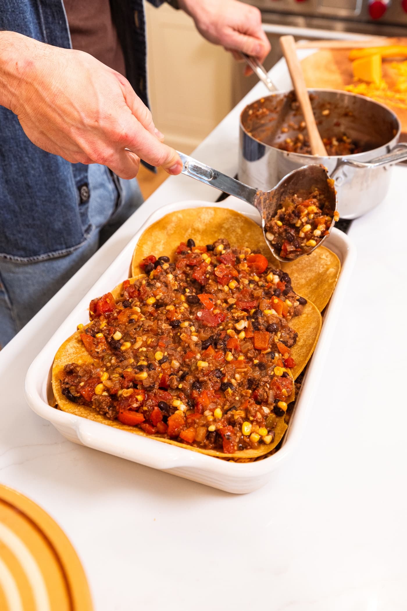 Ladle pouring ground beef mixture on top of corn tortillas in a rectangular baking dish with rest of the mixture to the side.
