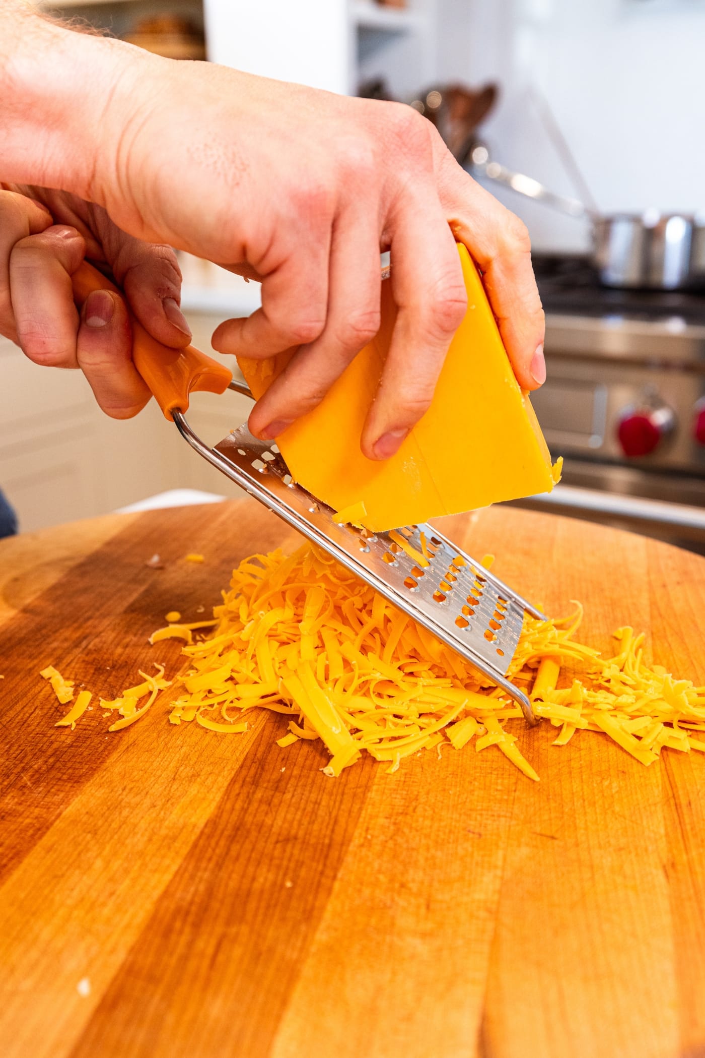 Hand using grater to grate cheese on a wooden cutting board into shreds.