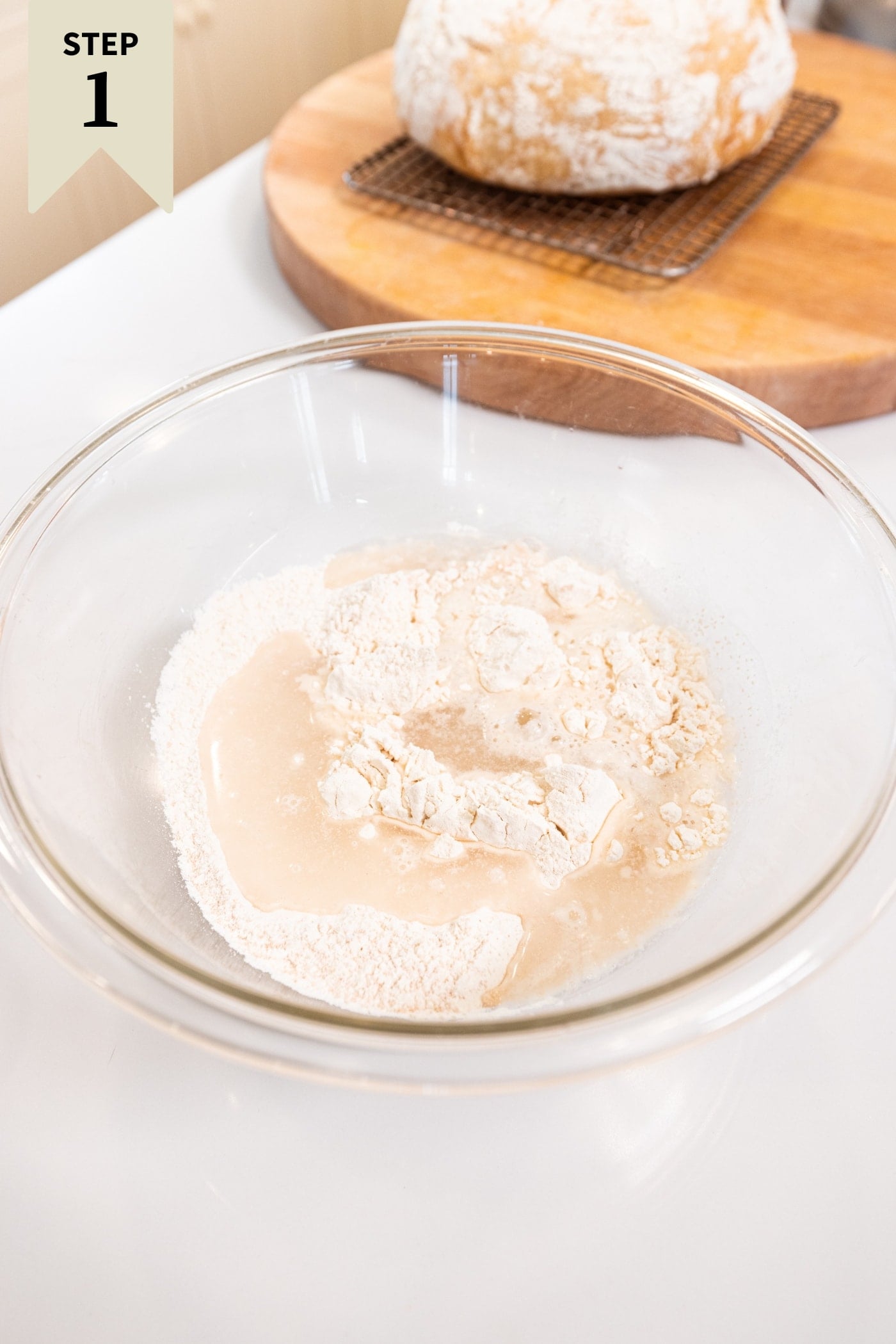 Glass bowl filled with ingredients needed for an overnight sourdough bread on white surface.