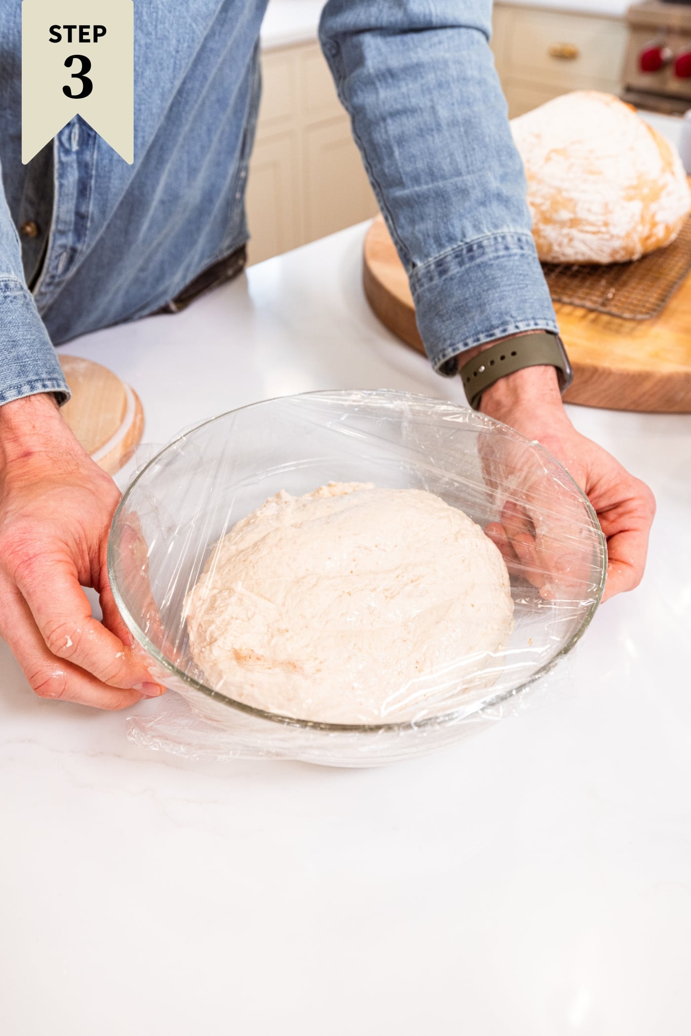 Glass bowl with hands placing plastic wrap over the top of a bowl with dough rising all on white surface.