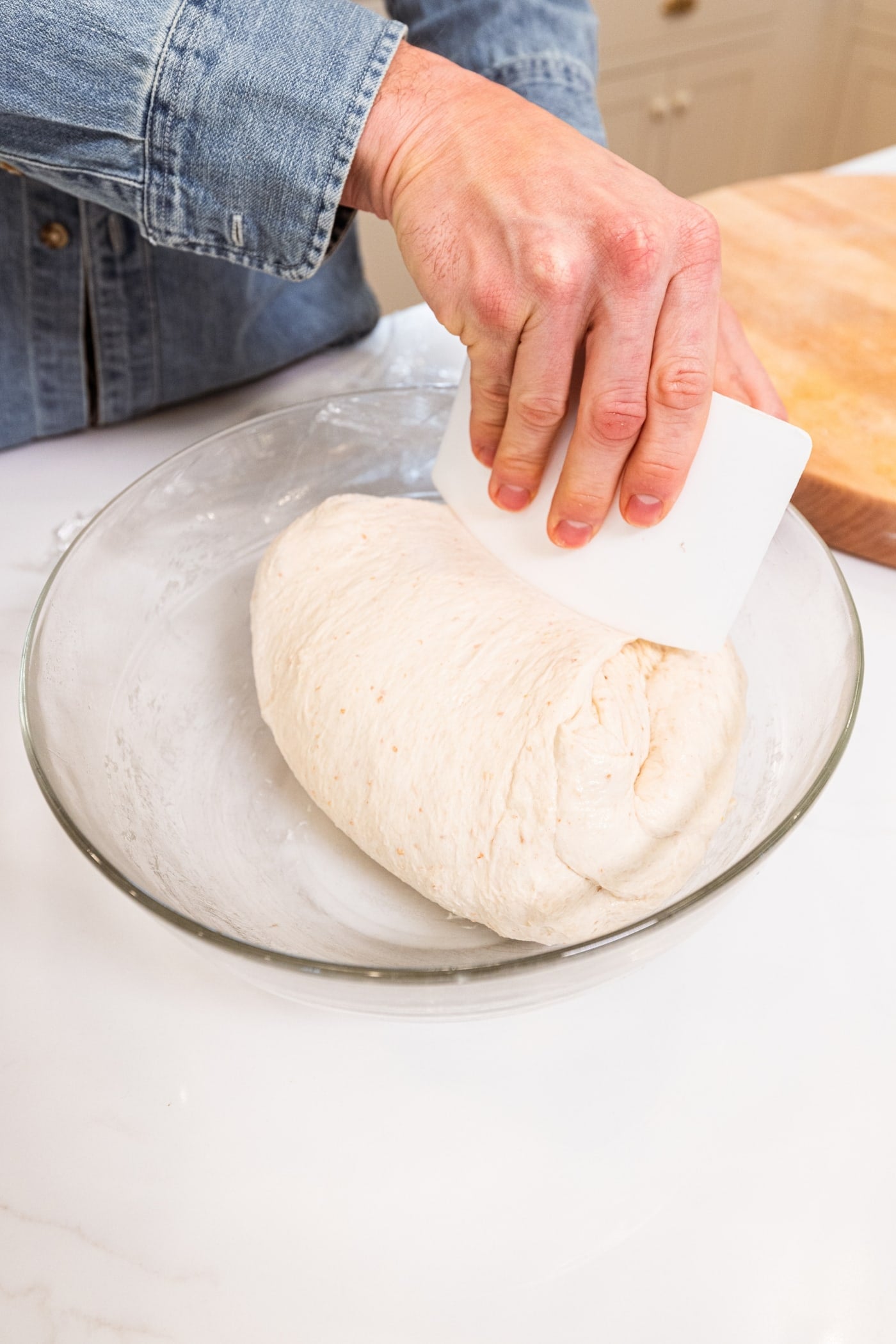 Hand using plastic scraper to turn dough over in a glass bowl on a white surface for overnight sourdough bread.