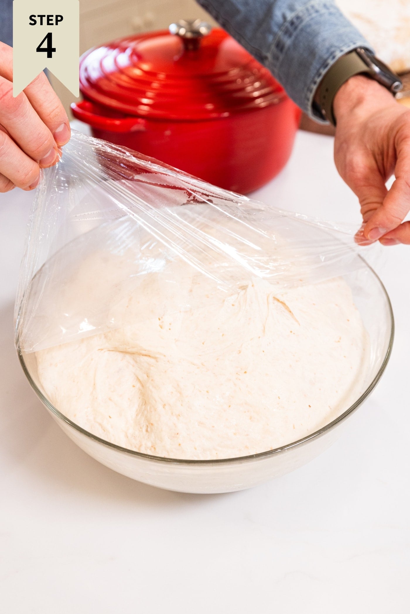 Plastic wrap being removed from a large glass bowl filled with risen sourdough.