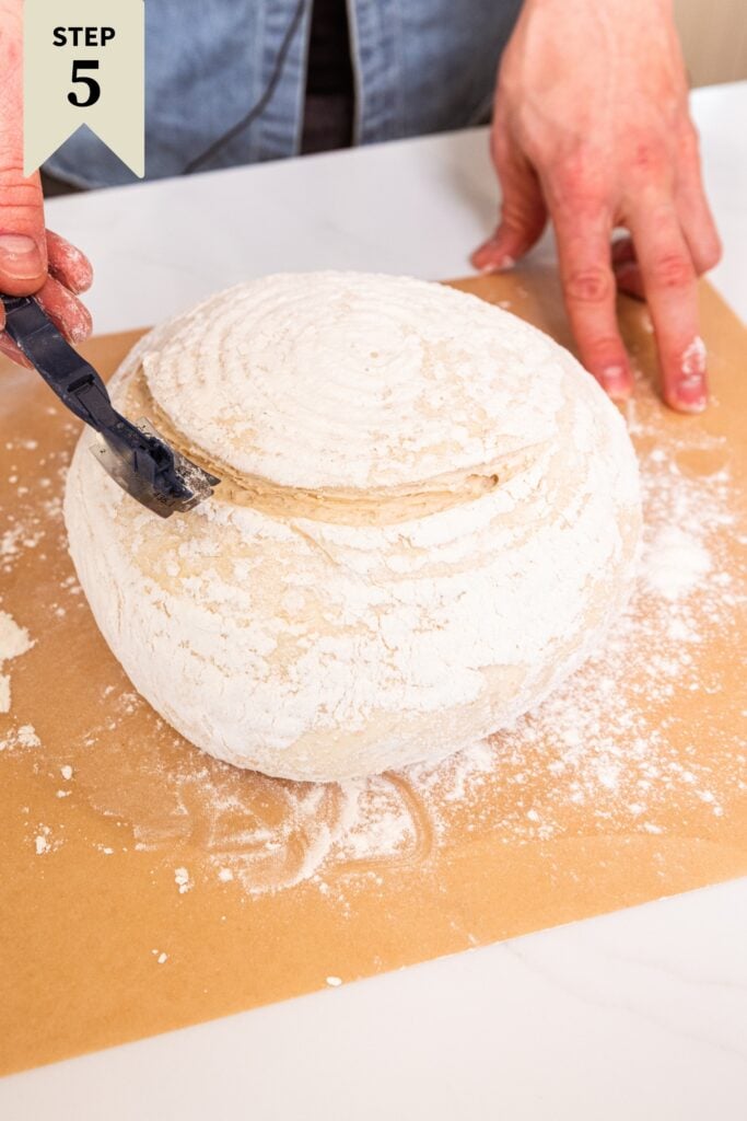 Hand using a lame to make an imprint on the top of an unbaked loaf of overnight sourdough bread.