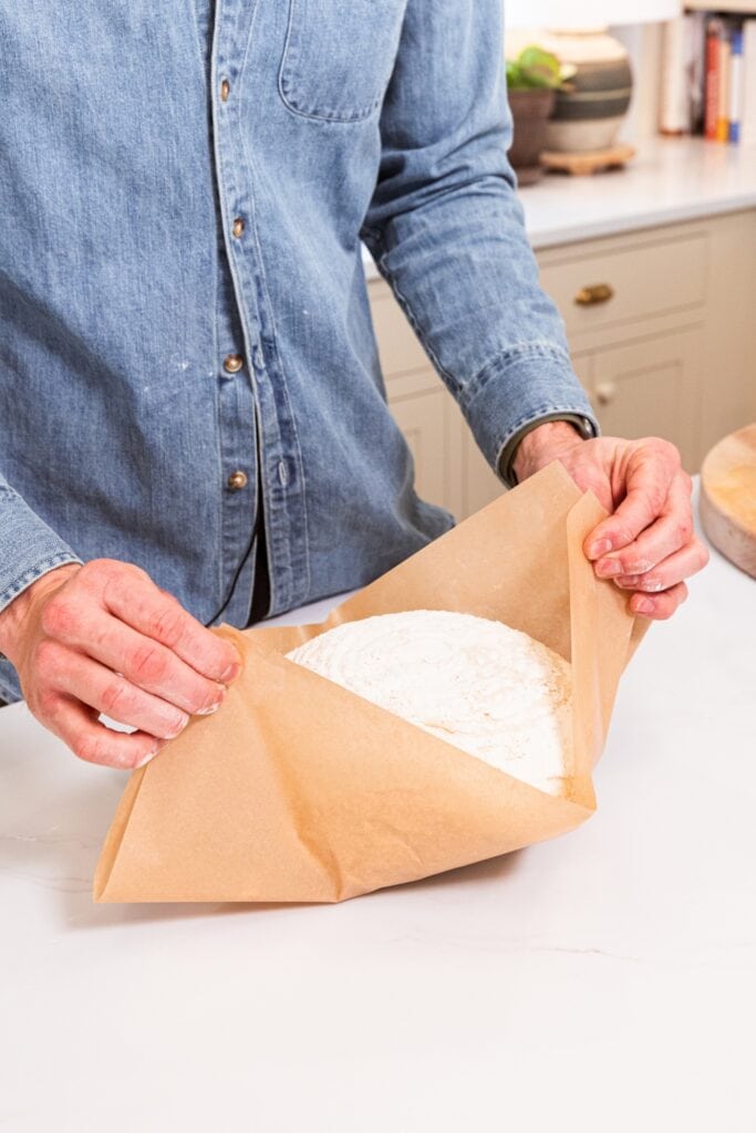 Two hands using a piece of parchment to pick up overnight sourdough bread from white surface.