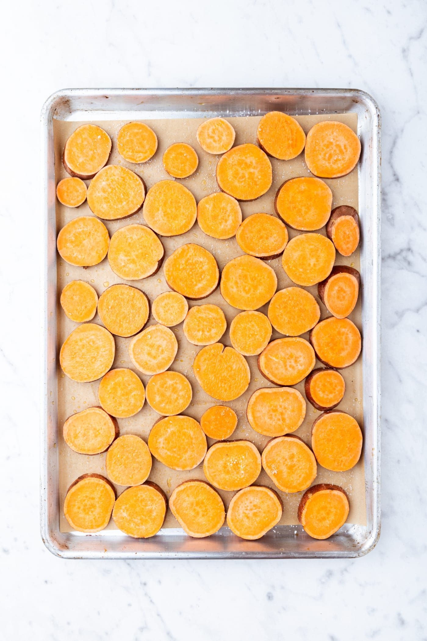 Sheet pan on marble surface with slices of sweet potato before being roasting in the oven.