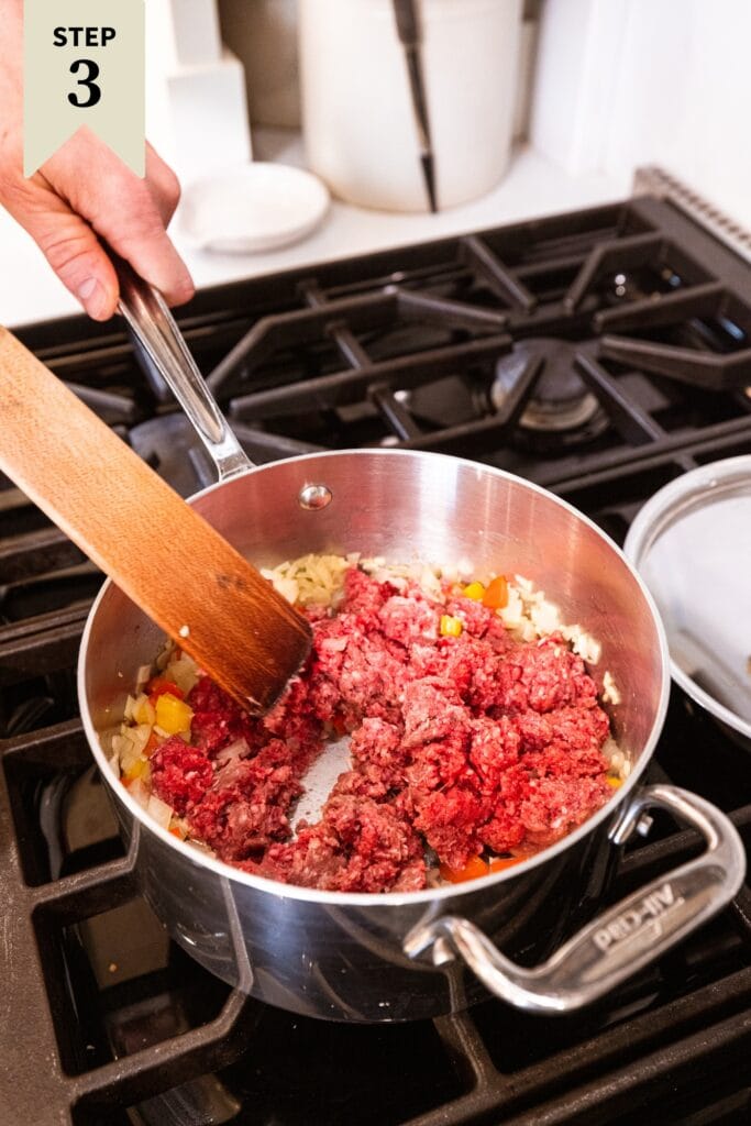 Raw ground beef in a metal pot on the stovetop with browned vegetables underneath.