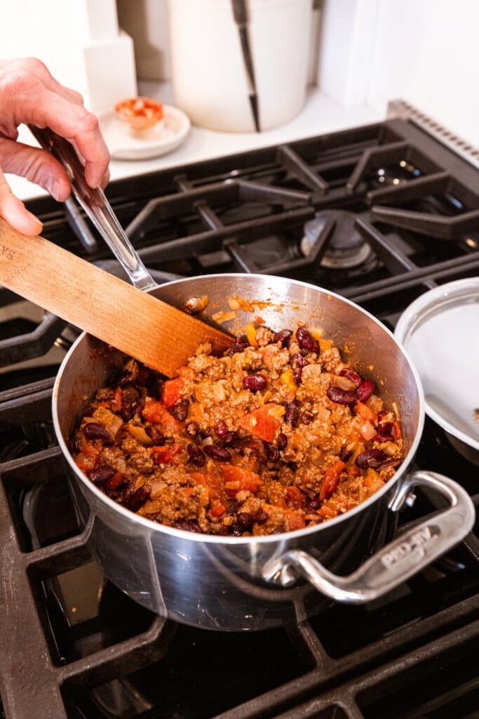 Hand using wooden spatula to mix together beans and tomatoes with cooked ground beef in a metal pot.