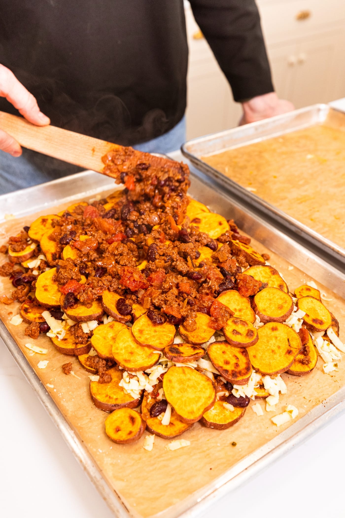 Hand using wooden spatula to spread chili on top of sweet potato slices on a parchment-lined baking sheet.