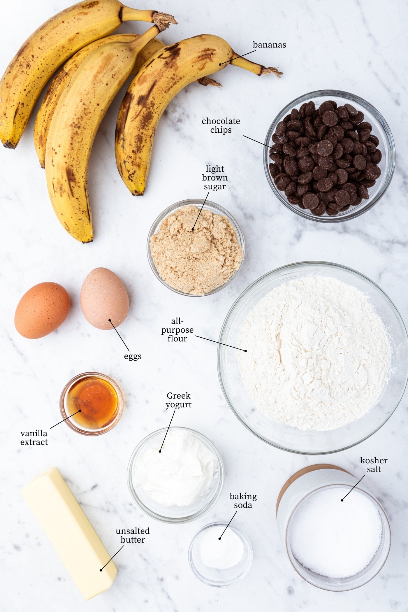 White marble surface filed with ingredients needed to prepare chocolate chip banana bread including bananas and chocolate chips.