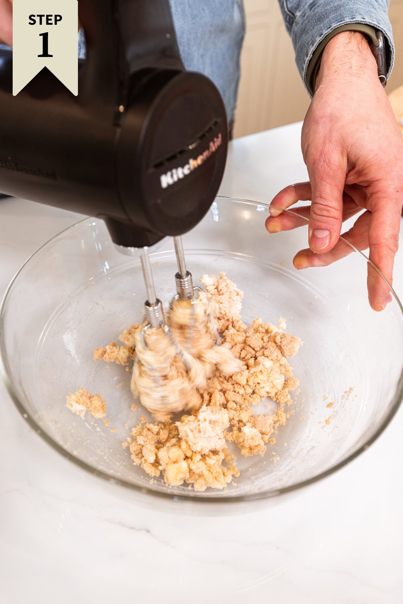 Glass bowl filled with sugar and butter being mixed together with hand mixer on white surface.
