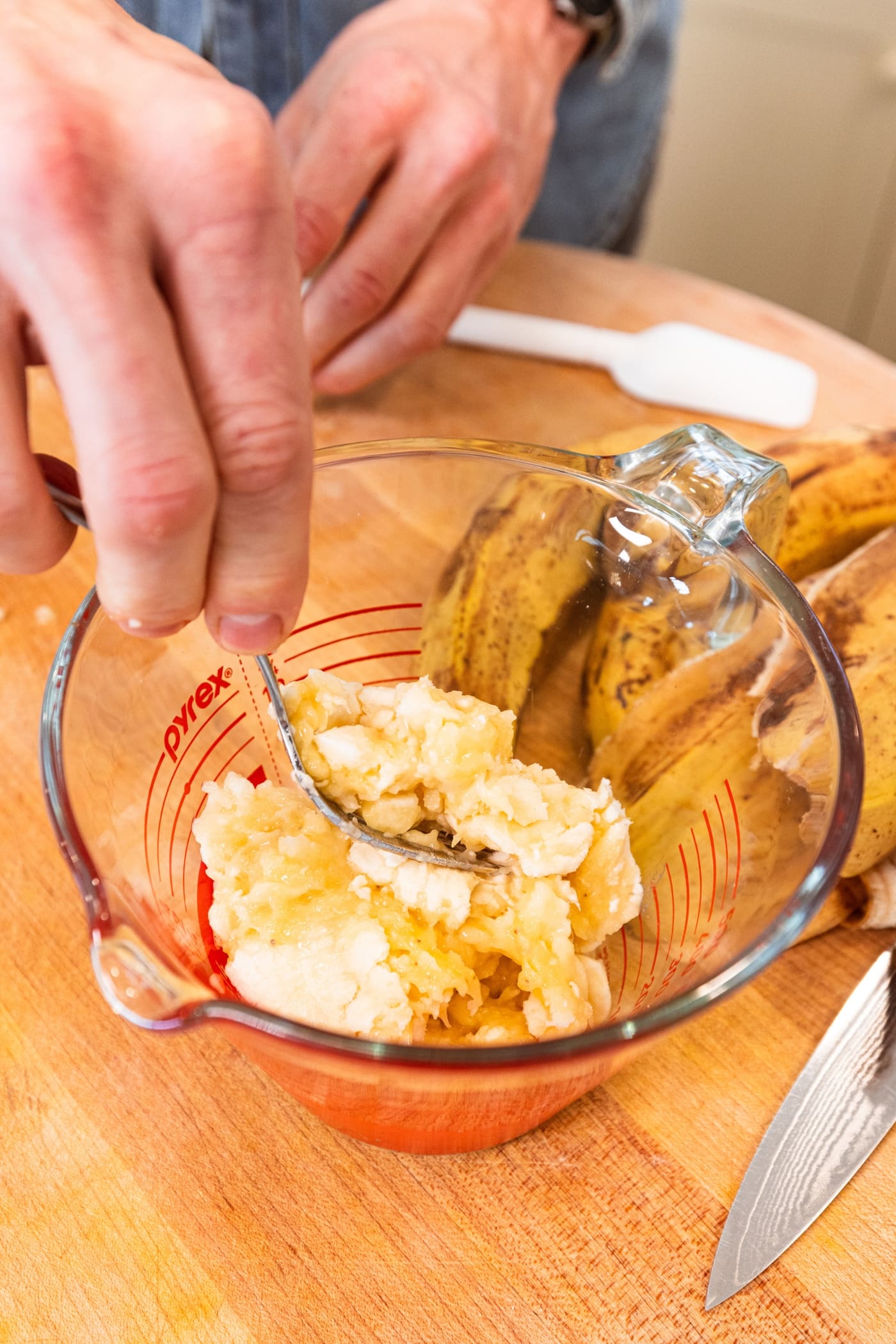 Hand using fork to place mashed banana pieces in a measuring cup on a wooden surface.