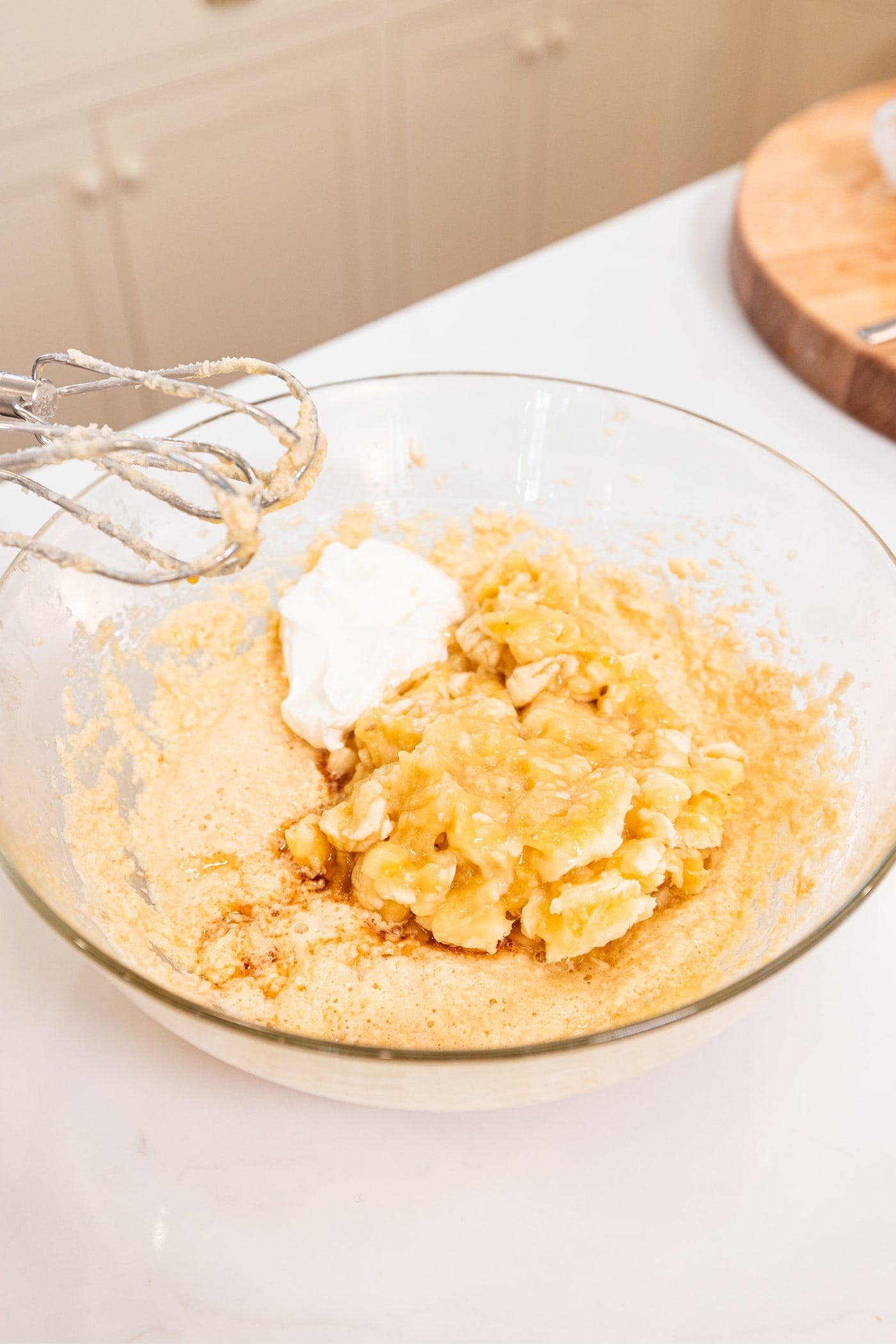 Glass bowl with mashed banana and yogurt along with sugar and butter mixture all on white surface.