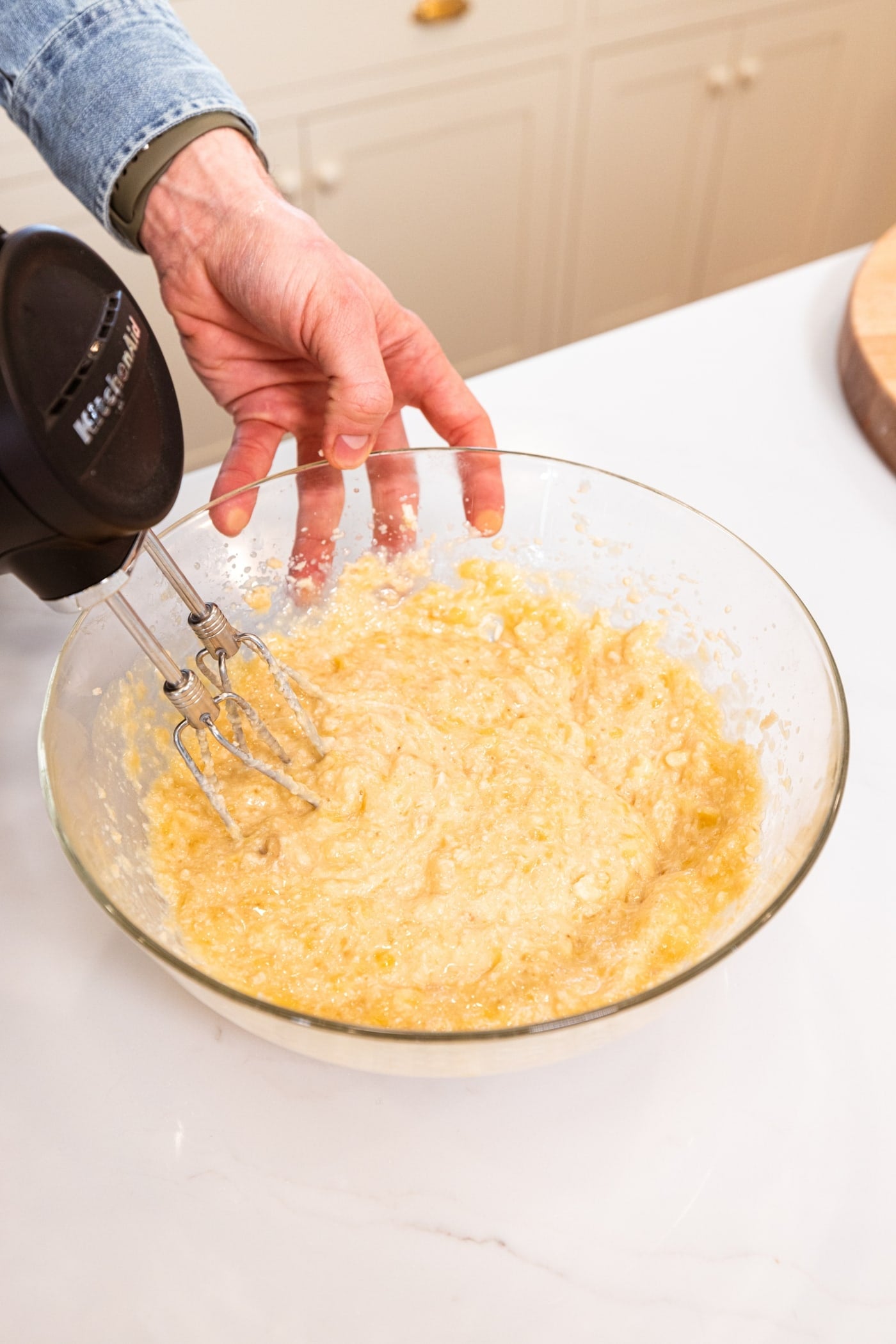 Hand mixer mixing together bananas with sugar mixture for chocolate chip banana bread all in glass bowl.