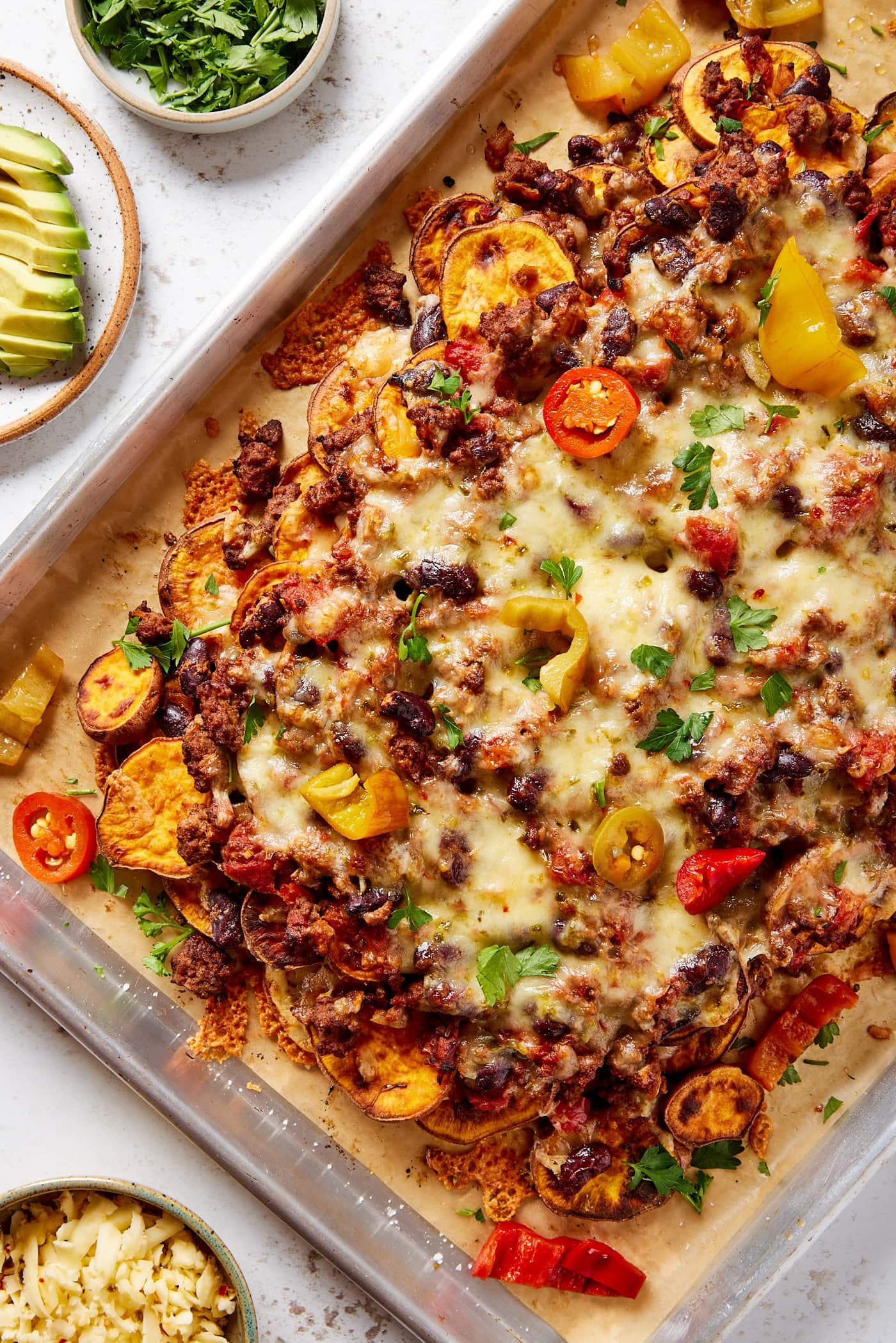 Metal sheet pan filled with sweet potato chili nachos with peppers, cilantro, and cheese sprinkled on top with avocado to the side.