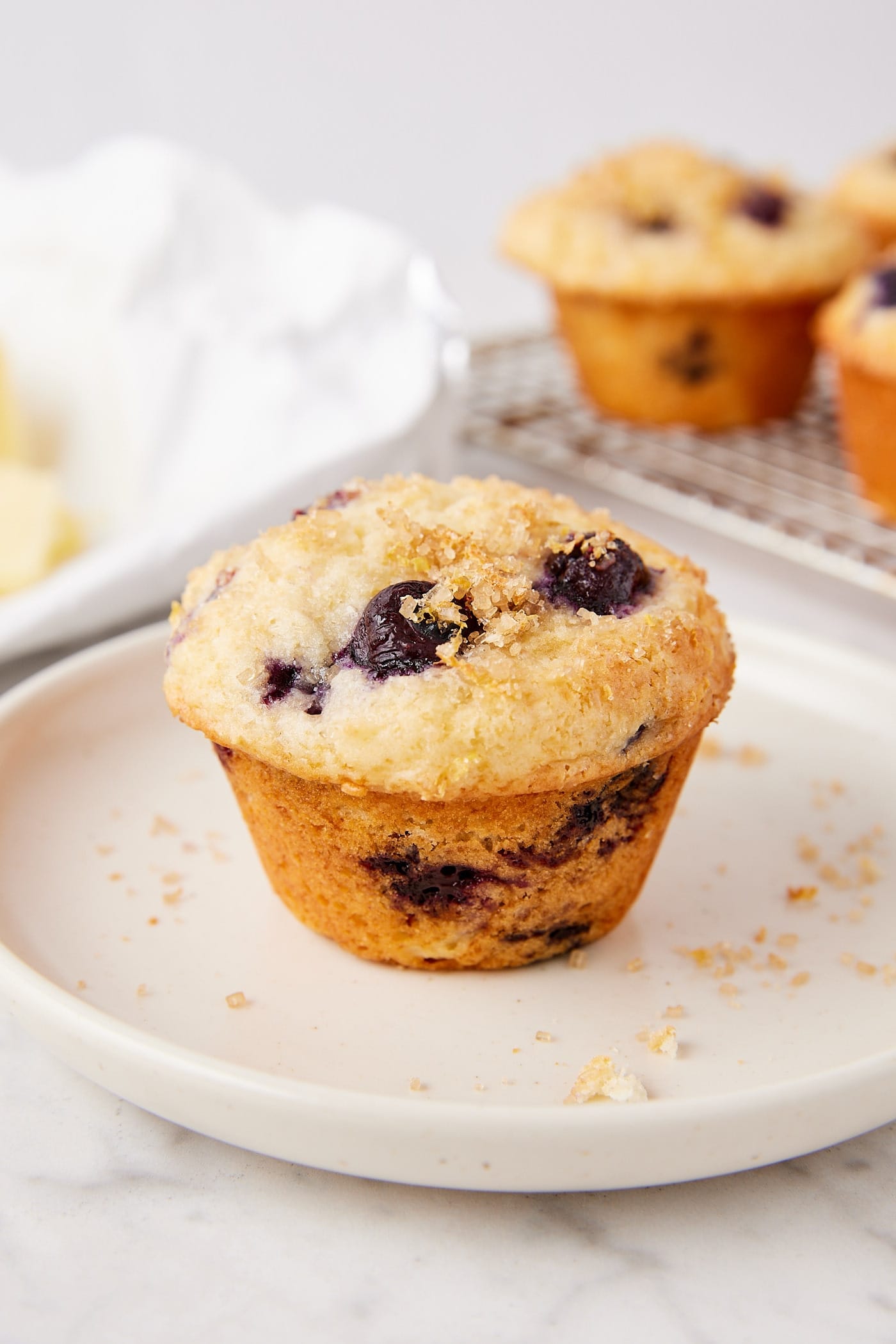 White plate with blueberry muffin with lemon zest turbinado sugar topping with butter in the background.