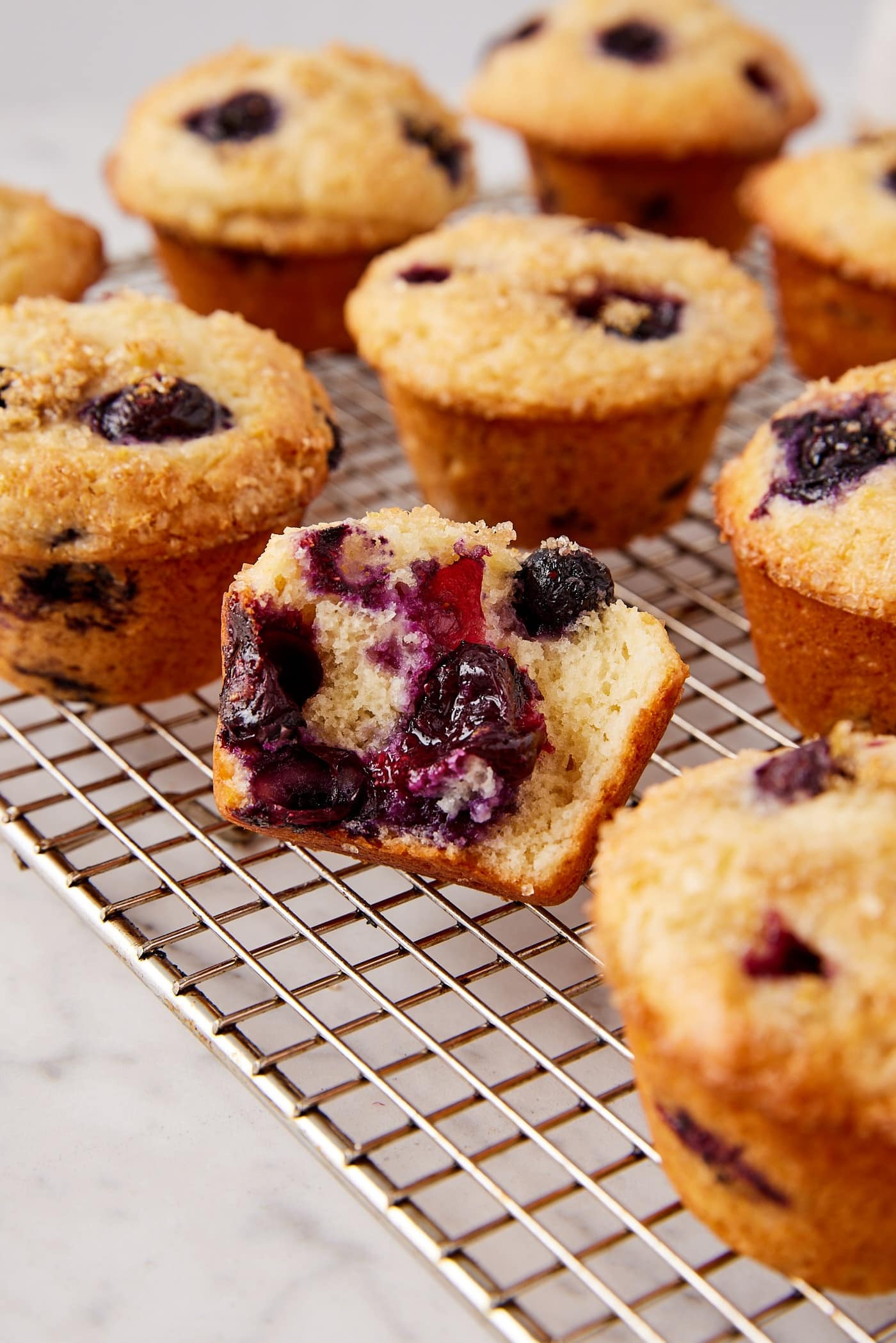 Marble surface with blueberry muffins cooling on a wire rack with one muffin torn in half showing interior texture.