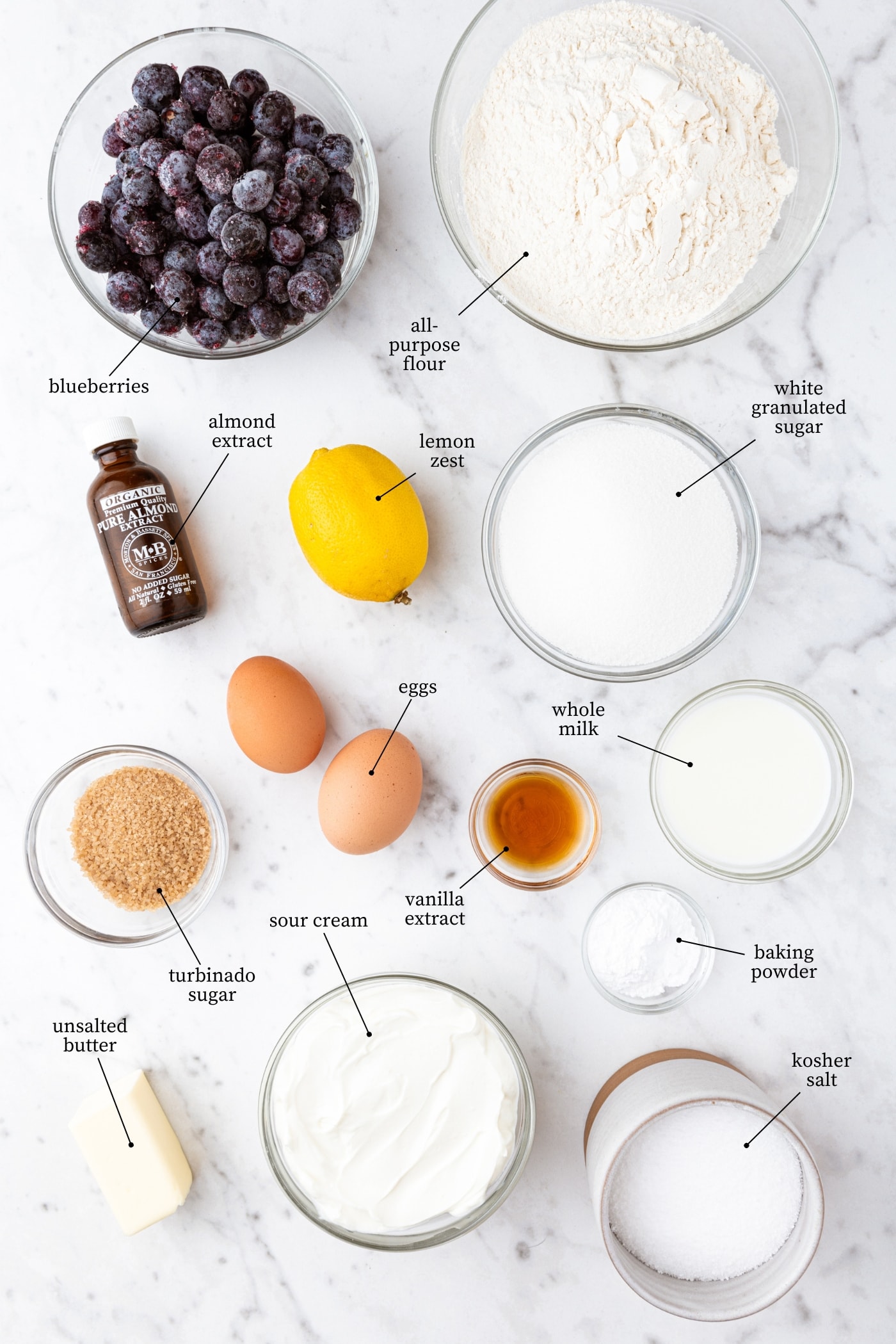White marble surface filled with all ingredients needed for blueberry muffins including flour, sugar, eggs, spices, blueberries, and more.