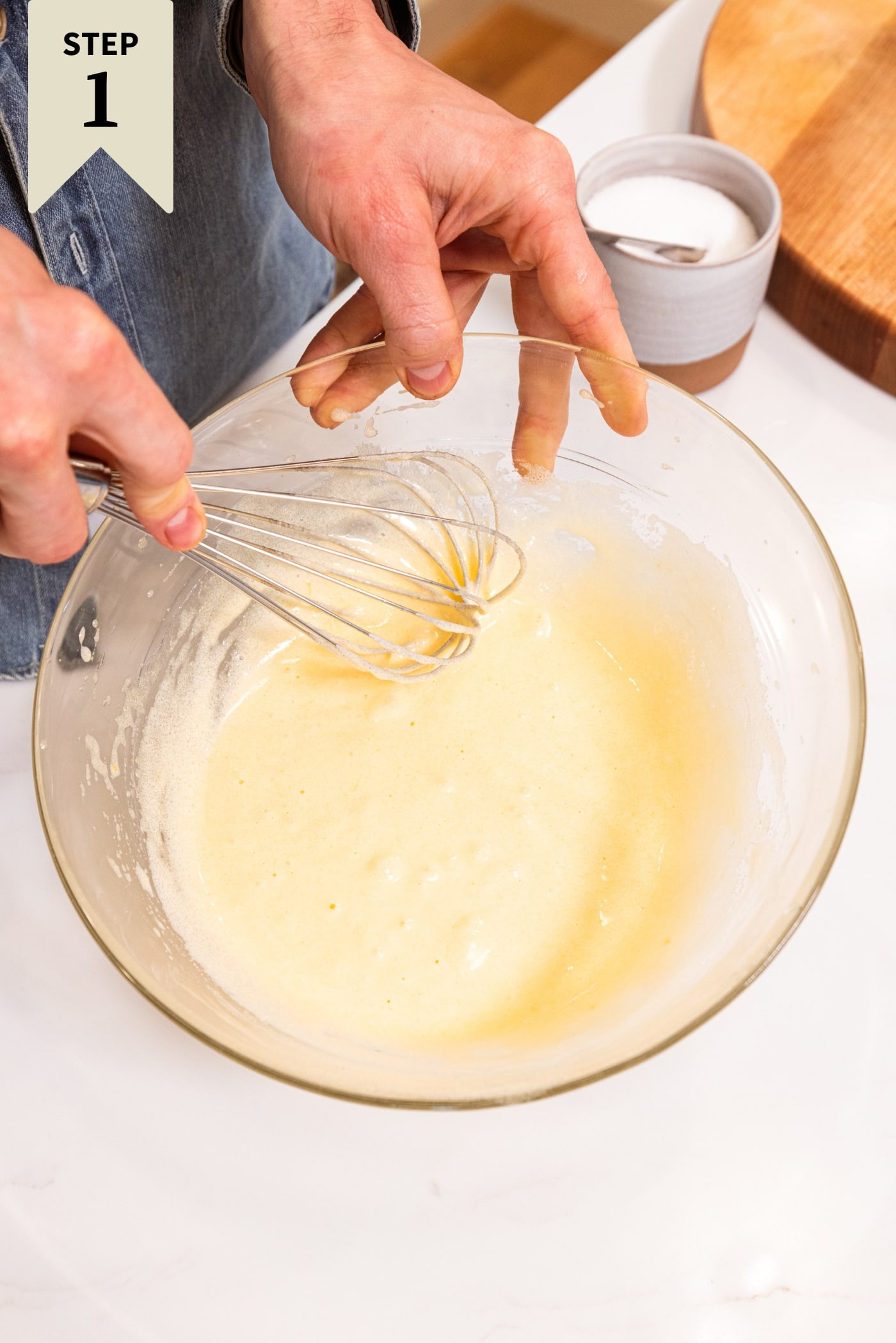 Glass bowl filled with batter for blueberry muffins with whisk whisking everything together all on white surface.