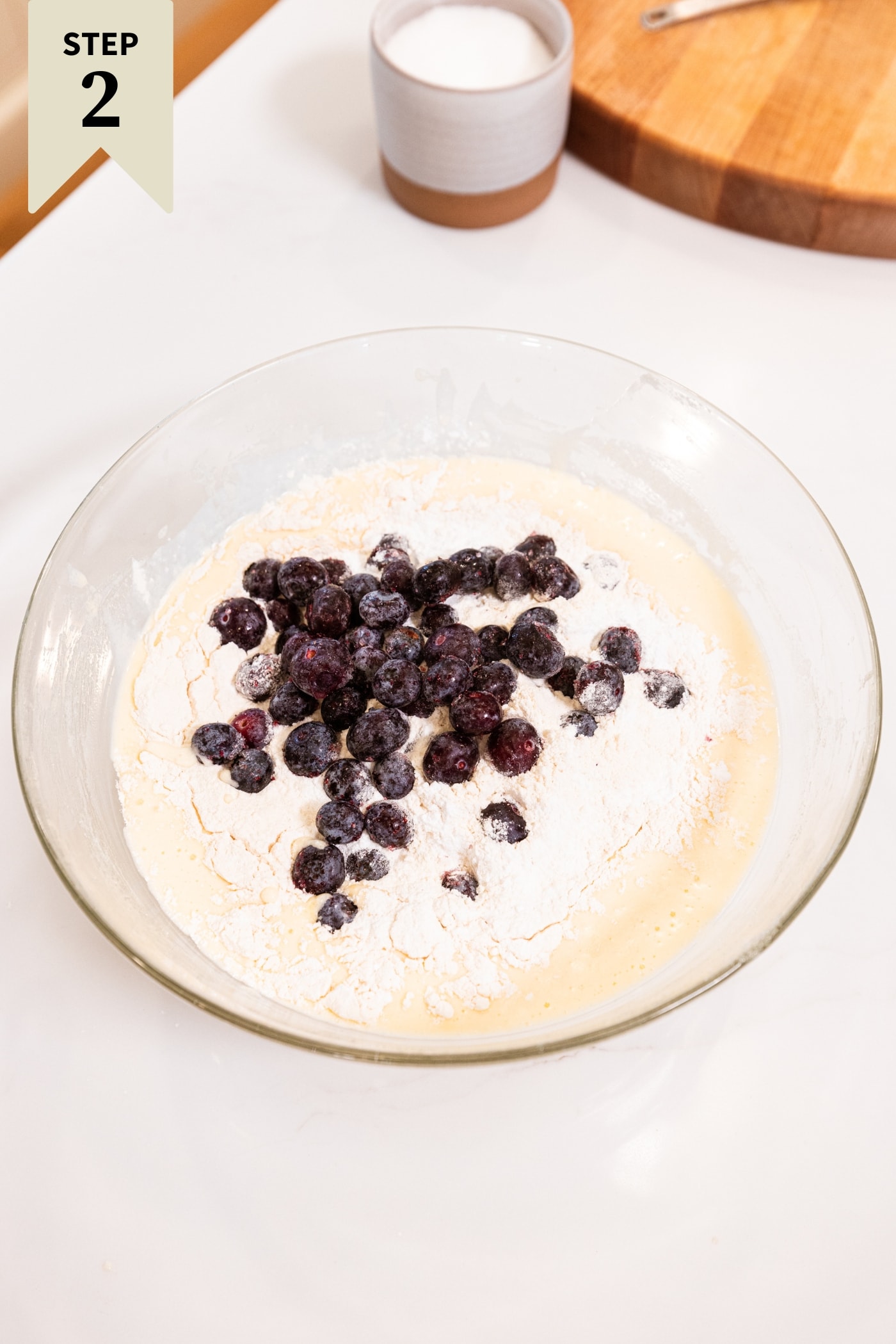 Glass bowl filled with flour on top of batter with frozen blueberries sprinkled on top all for blueberry muffins.