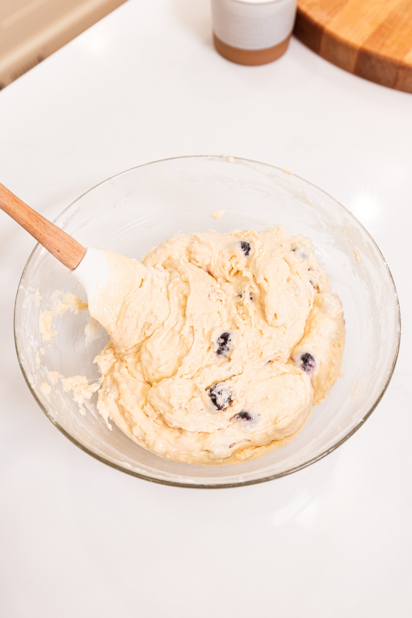 Glass bowl filled with batter with blueberries folded in all on white surface.